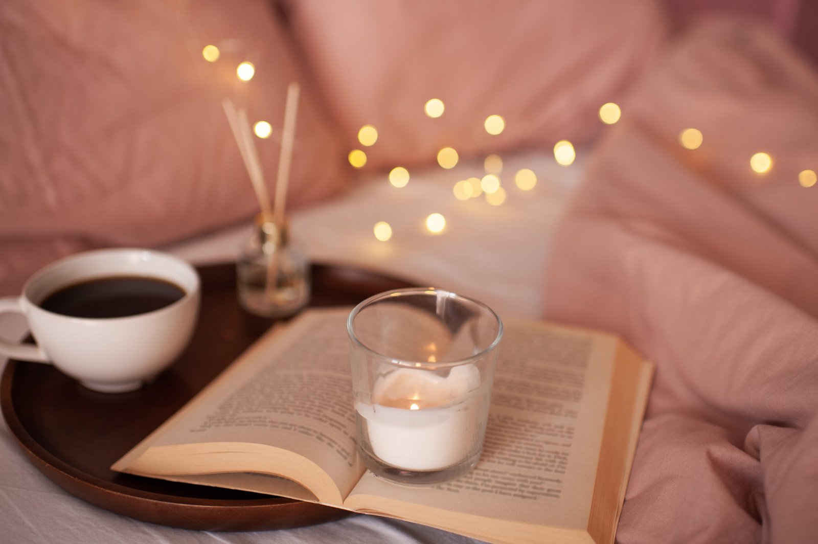 Burning candle staying on open paper book and cup of tea with aroma sticks at background over glowing lights. Cozy atmosphere concept. 