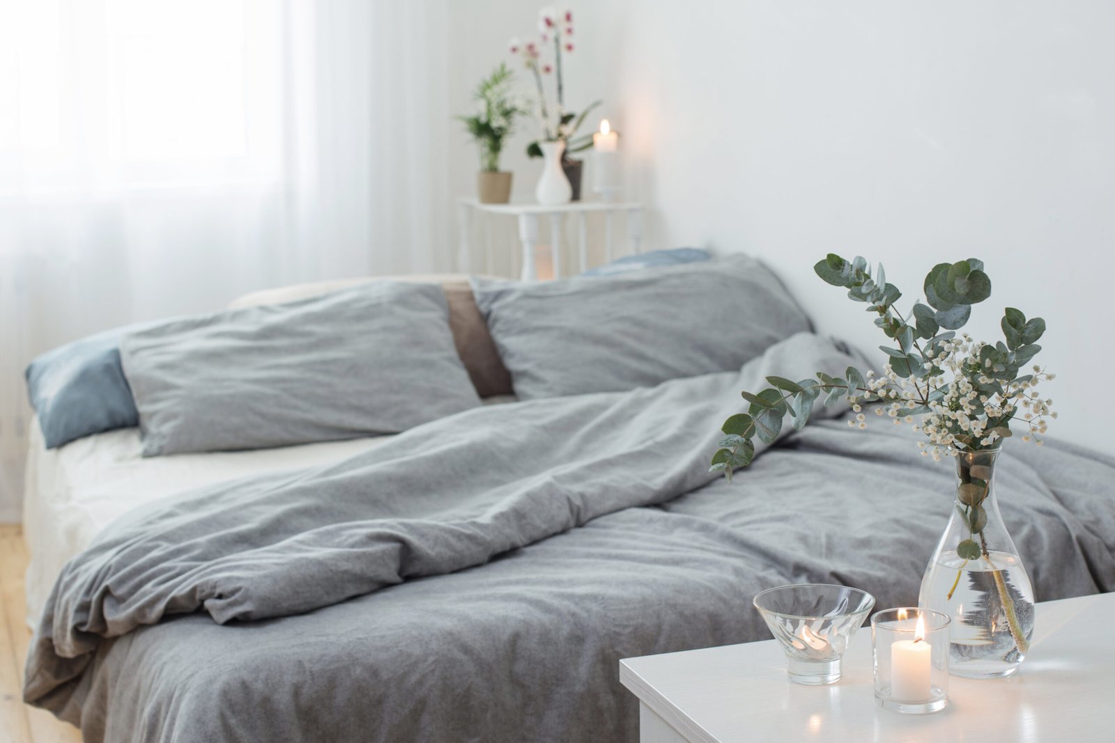 Burning candles and eucalyptus in glass vase in white bedroom, gray bed sheets.