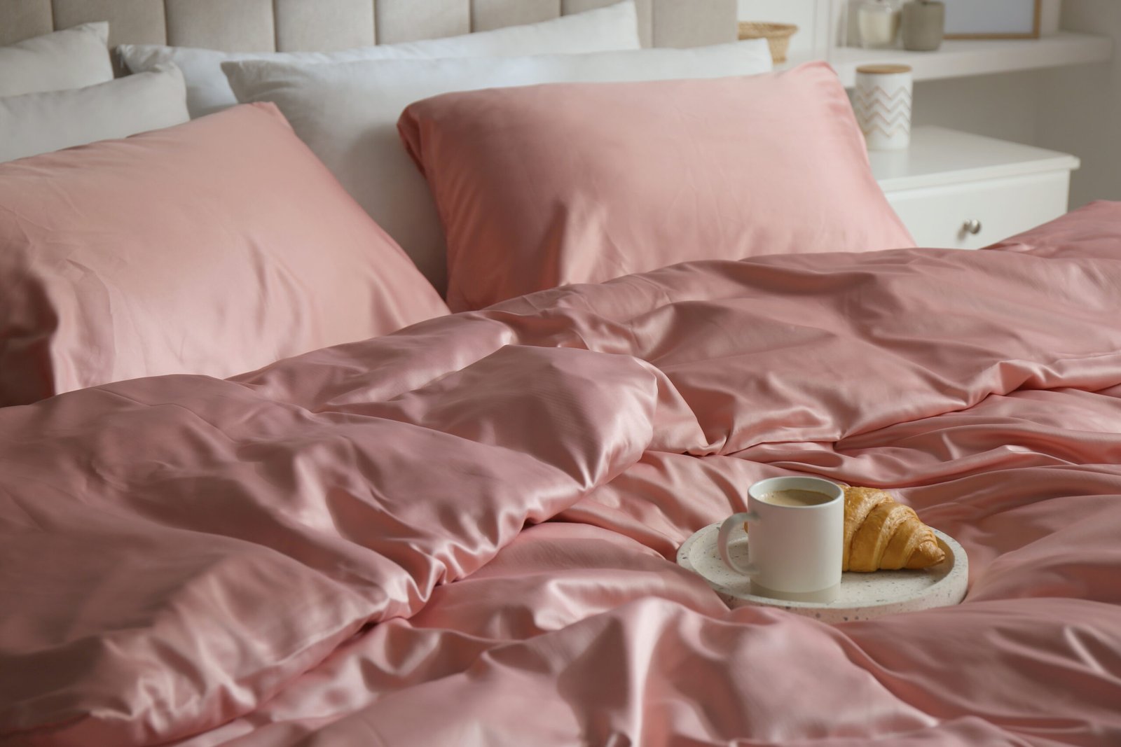 Croissant and coffee in breakfast tray on bed with beautiful pink silk linens bed sheet.
