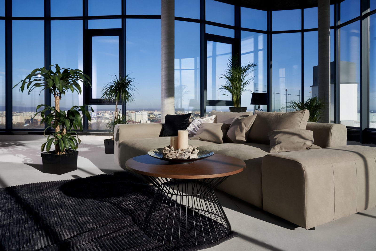 Front view of fashionable interior spacious room with large panoramic window with incredible view on city and cozy grey sofa with pillows and green flowerpots.