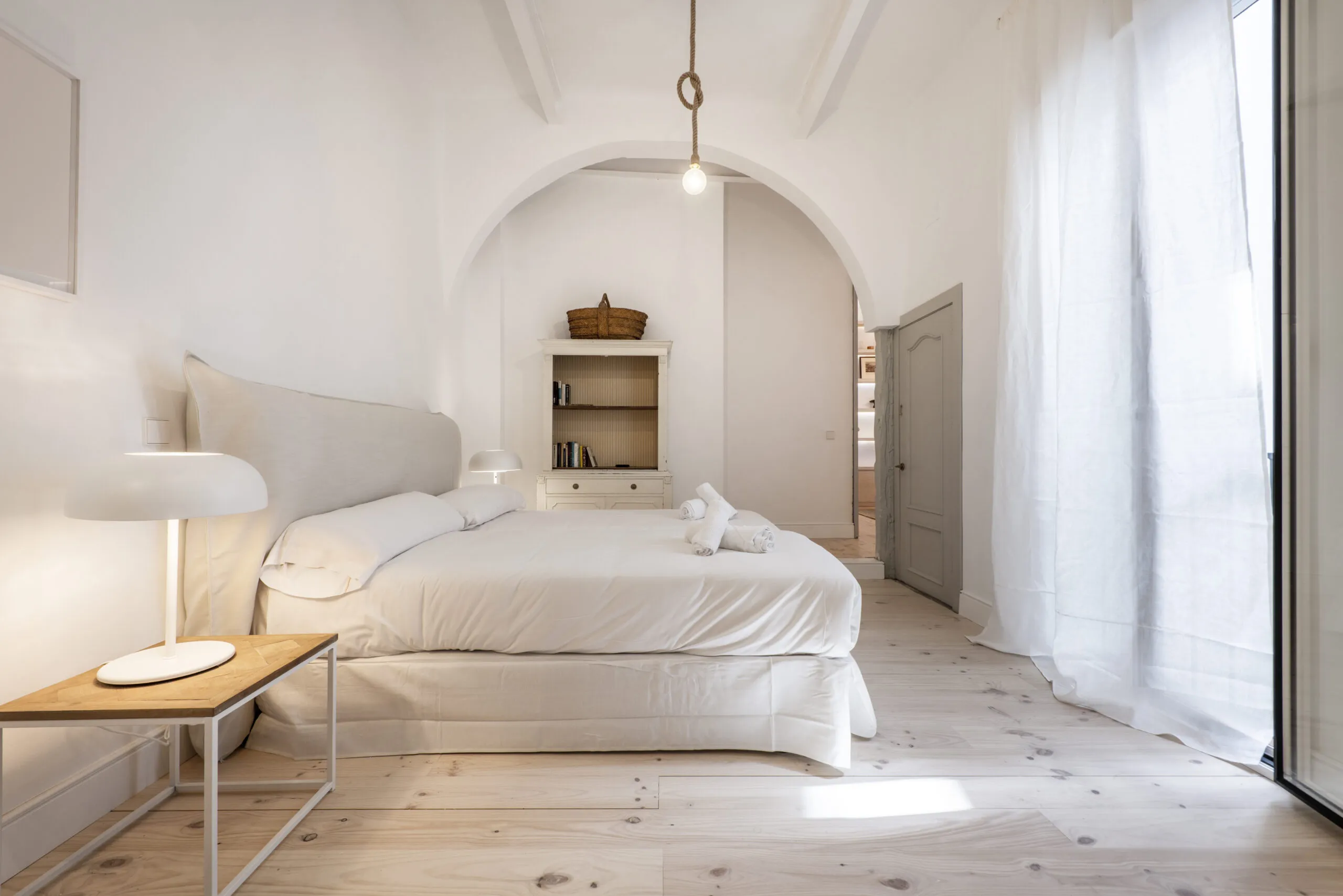 Nice bedroom with a king size bed, light pine wood floors, white curtains and a semicircular chest on one side of a room.