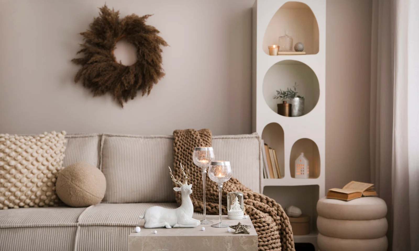 Amazing composition on white design shelf with christmas decoration, lights, gifts, lanterns, deer, candles, stars, round wreath white corduroy sofa and pouf.