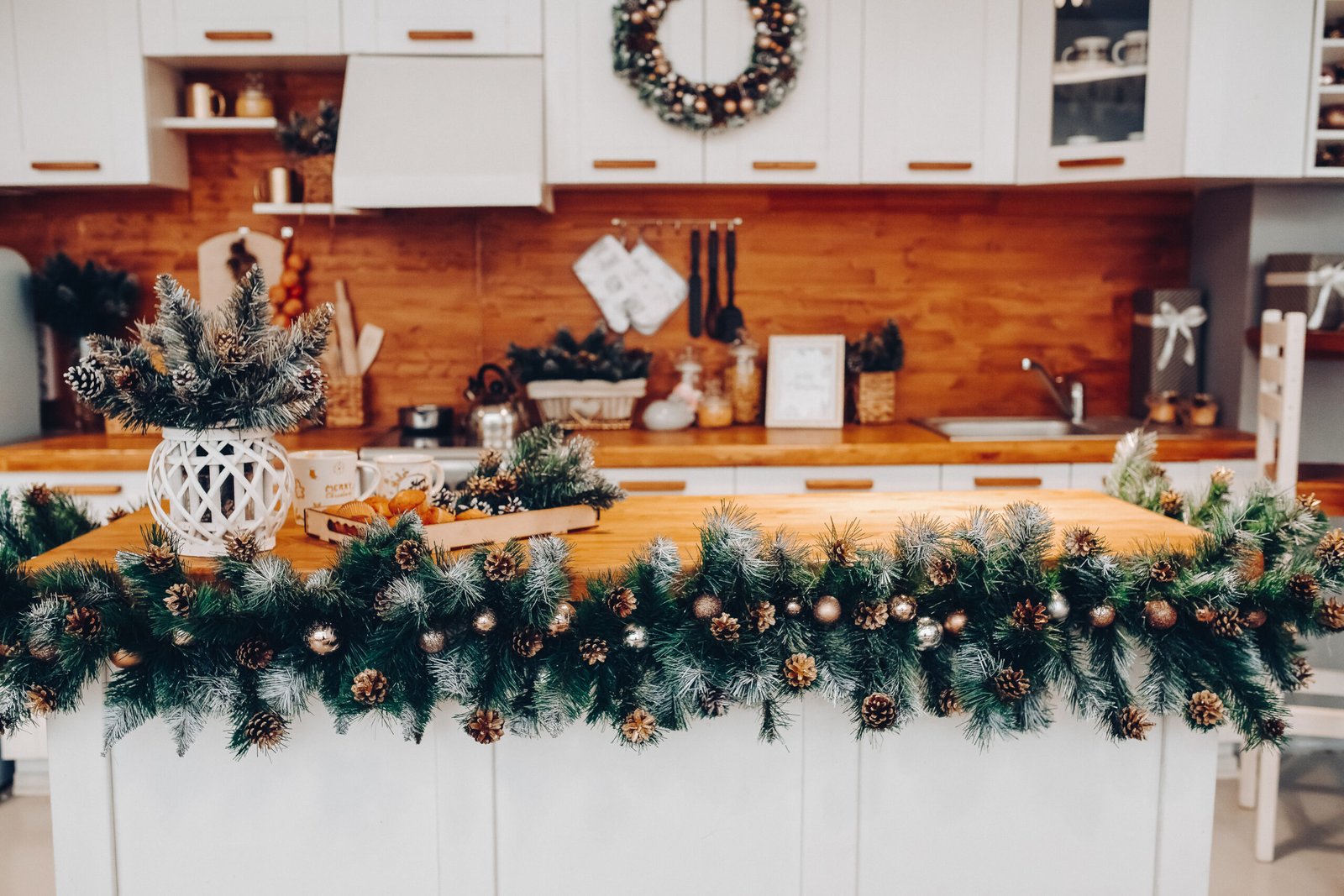 View over beautiful white kitchen with Christmas decorations all over cupboards and kitchen board. There is Christmas wreath on the cupboard. Natural fir tree branches with pine cones.