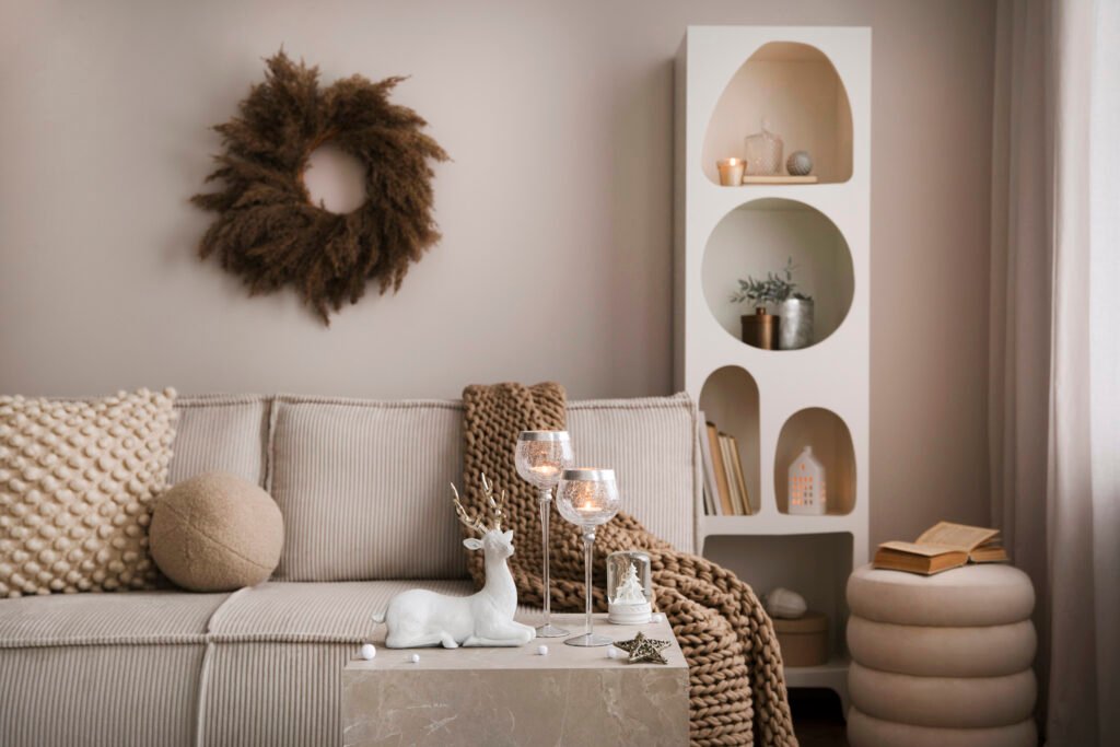 Amazing composition on white design shelf with christmas decoration, lights, gifts, lanterns, deer, candles, stars, round wreath white corduroy sofa and pouf.