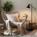 Scandi bedroom with a neutral bed, an armchair in a natural material and a black floor lamp. Next to the chair is stacked books.