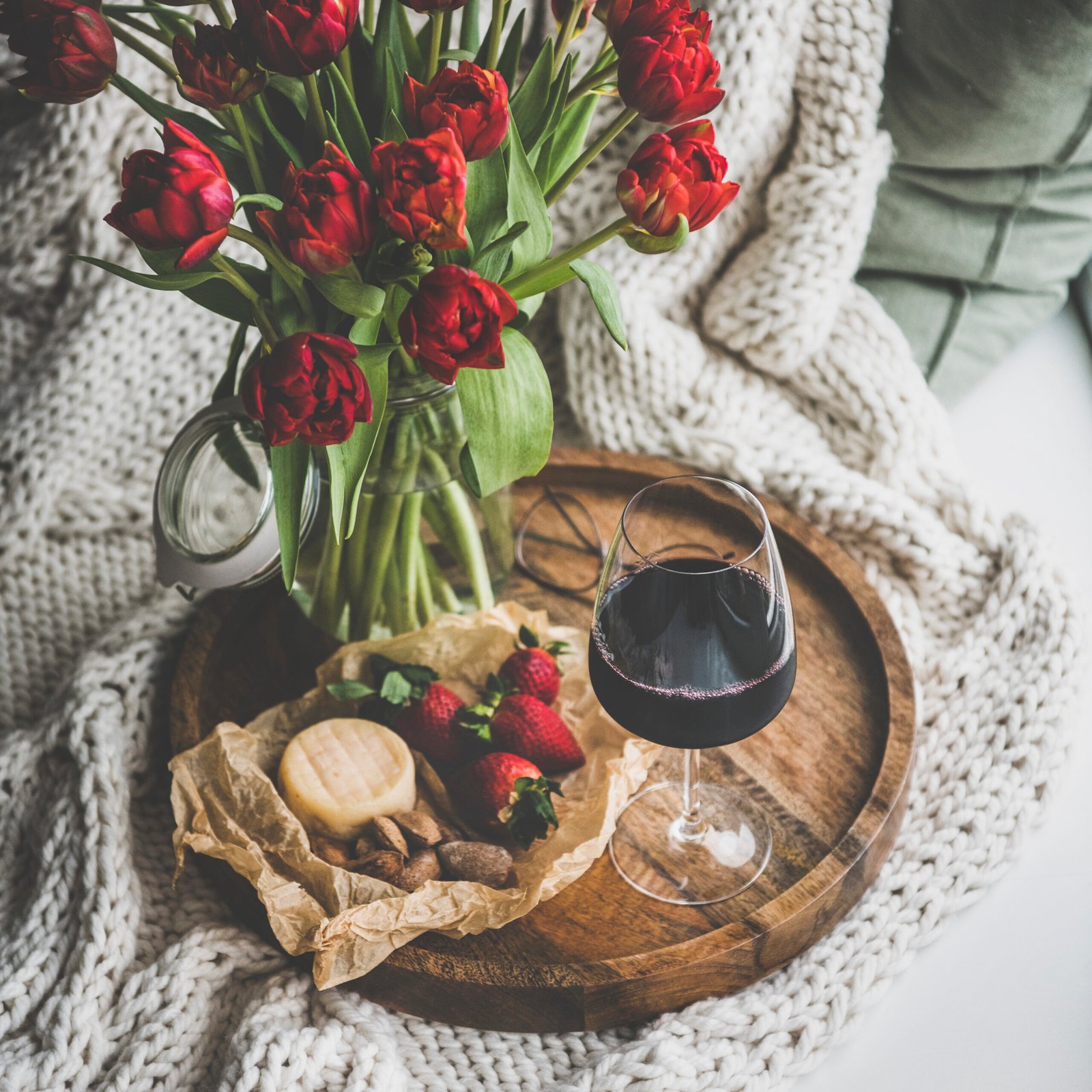 Wine snack set with flowers. Glass of red wine, cheese, roasted almonds, strawberries and bouquet of Spring red tulips on wooden tray over white knitted blanket, square crop. 