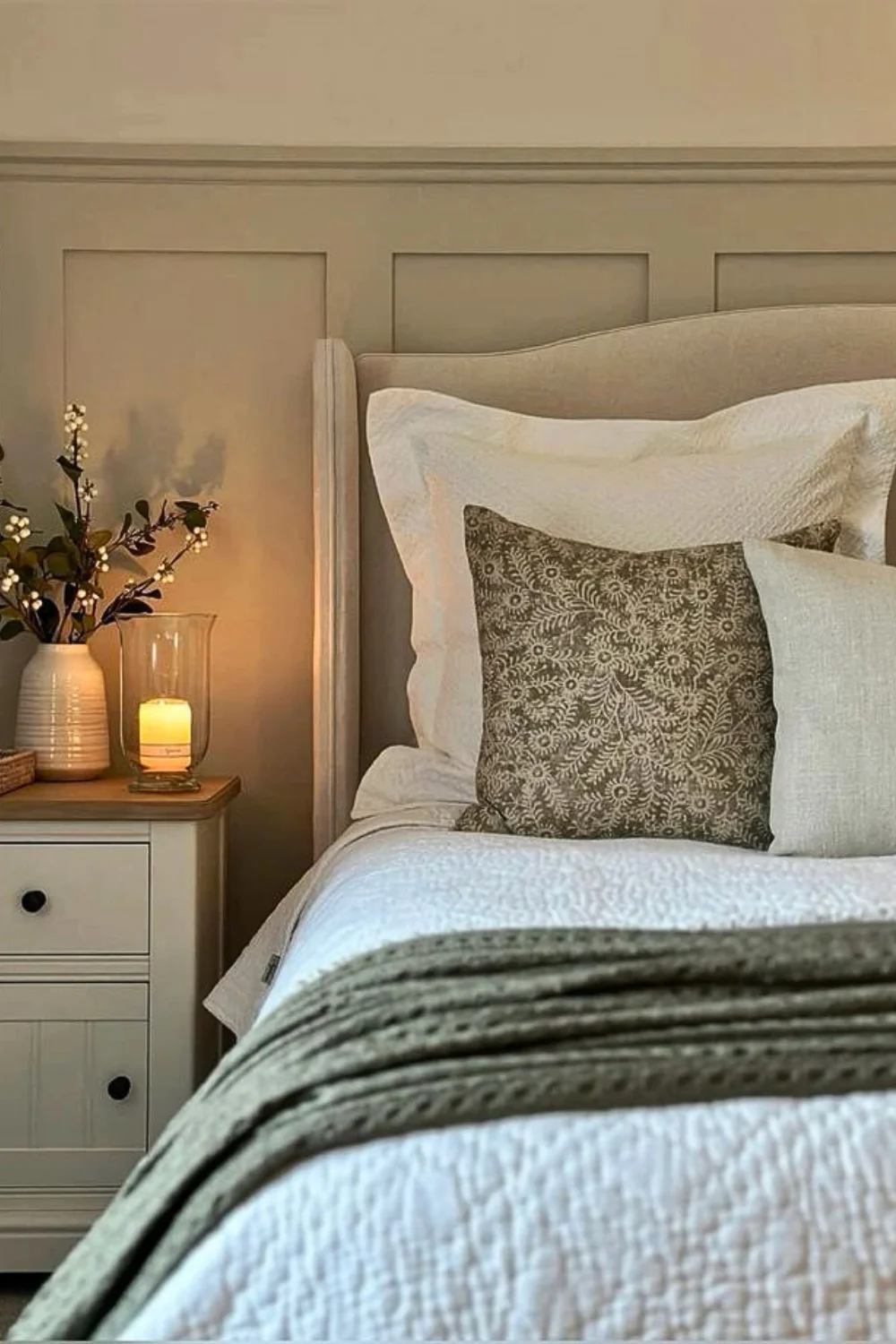 Cozy cottage style bedroom with a neutral bed frame and neutral bedding. A bedside table with a flower. 