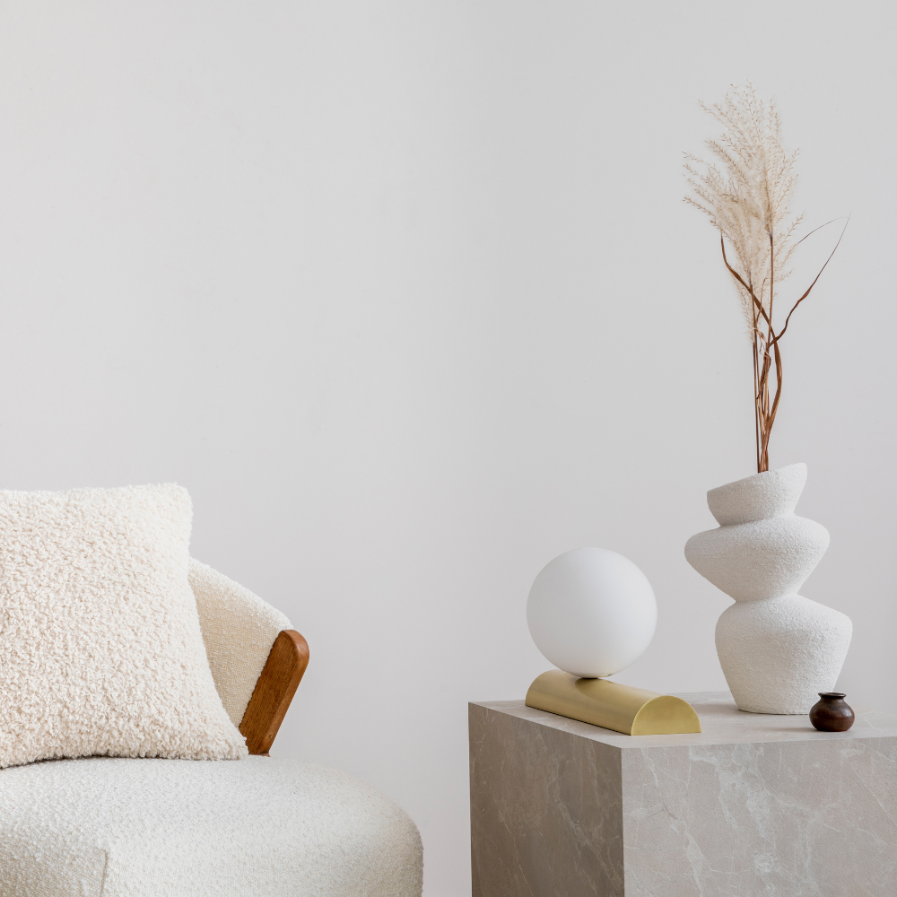 White chair with a white cozy pillow and a white wall behind it. A side table with a white vase on it. 