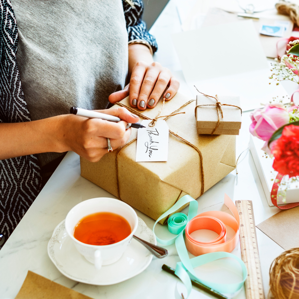 Female hands wrapping a gift with brown paper. A cup of tea and wrapping material.