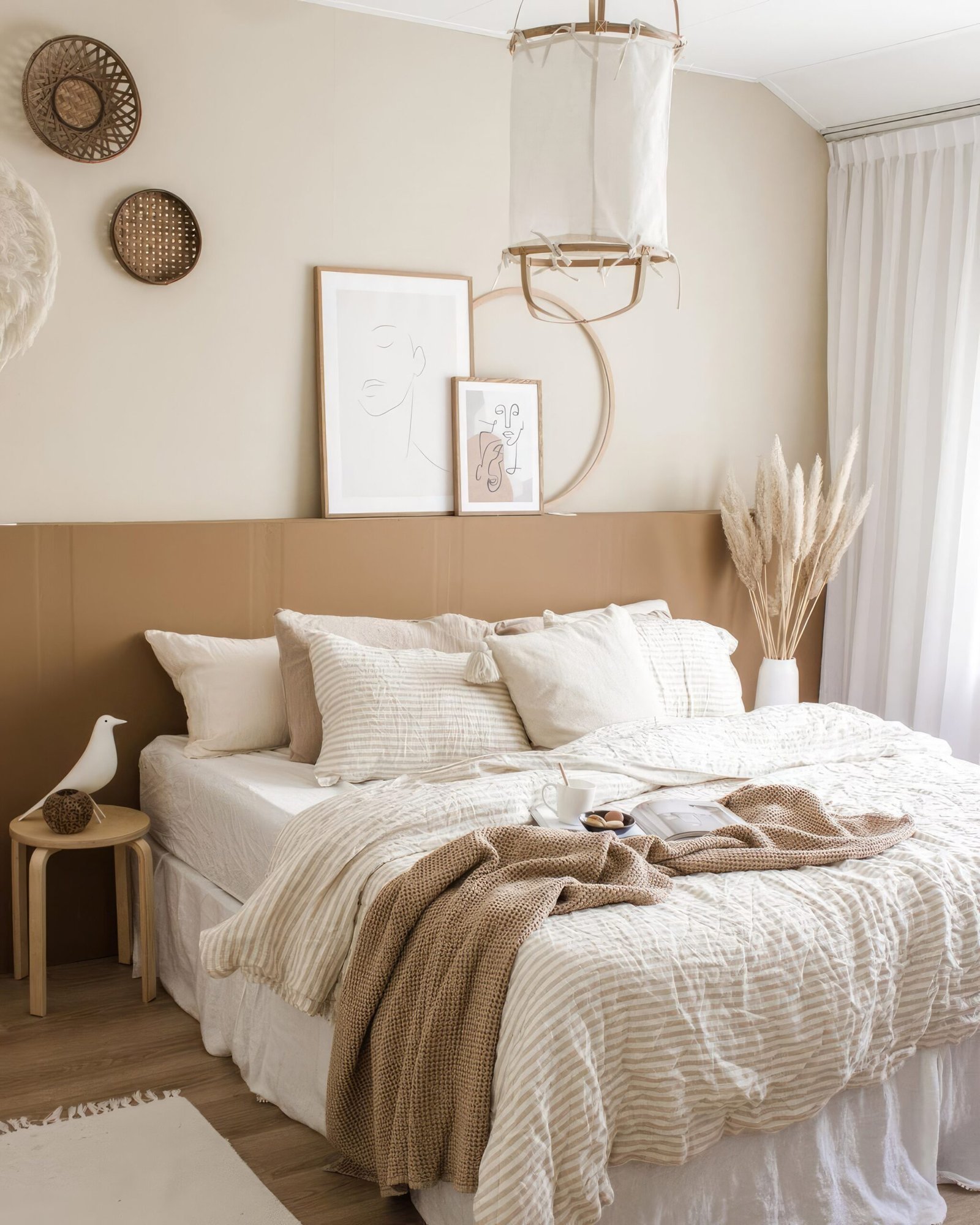 A bed frame with white bedding, a knit throw blanket, a wooden stool next to the bed. Pampas grass in a vase and wall art. Scaled.