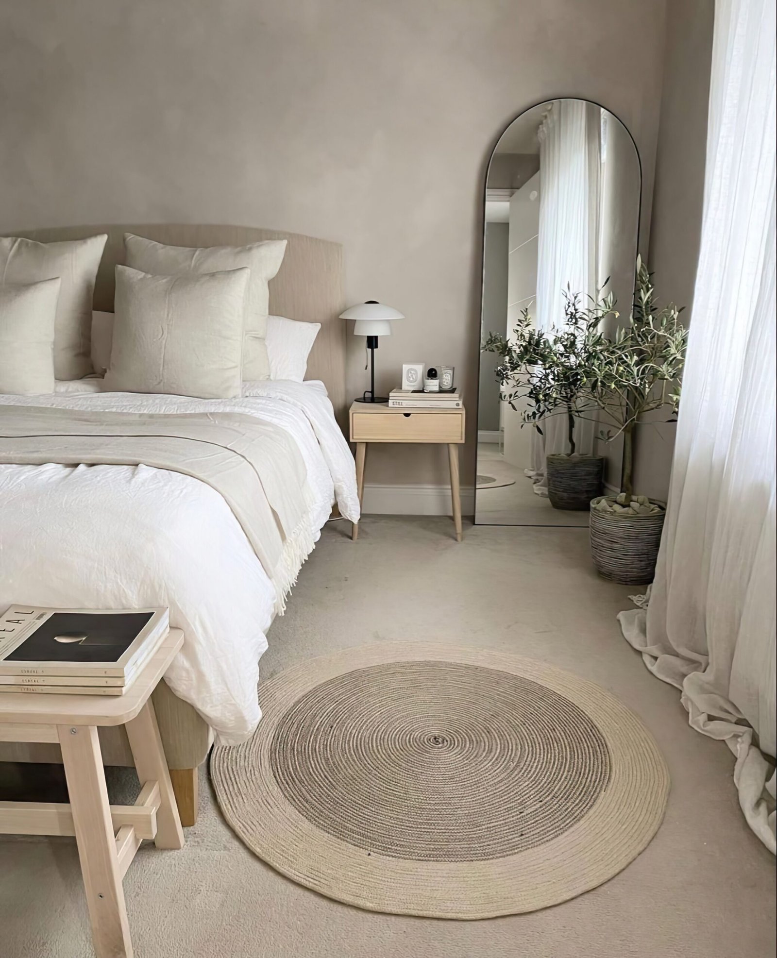 Bedroom with a large neutral bed, a mirror and a plant, a wooden bedside table and a rug.