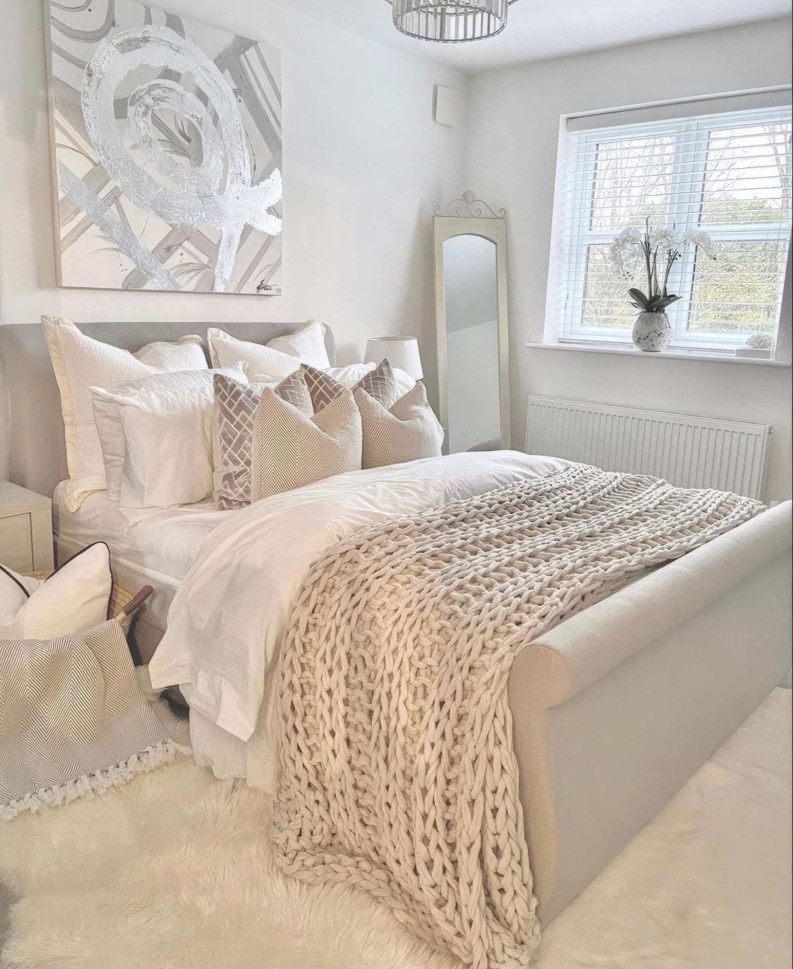 Bedroom with neutral bedding and a chunky thrown knit blanket. A large window and a mirror. A wall art.