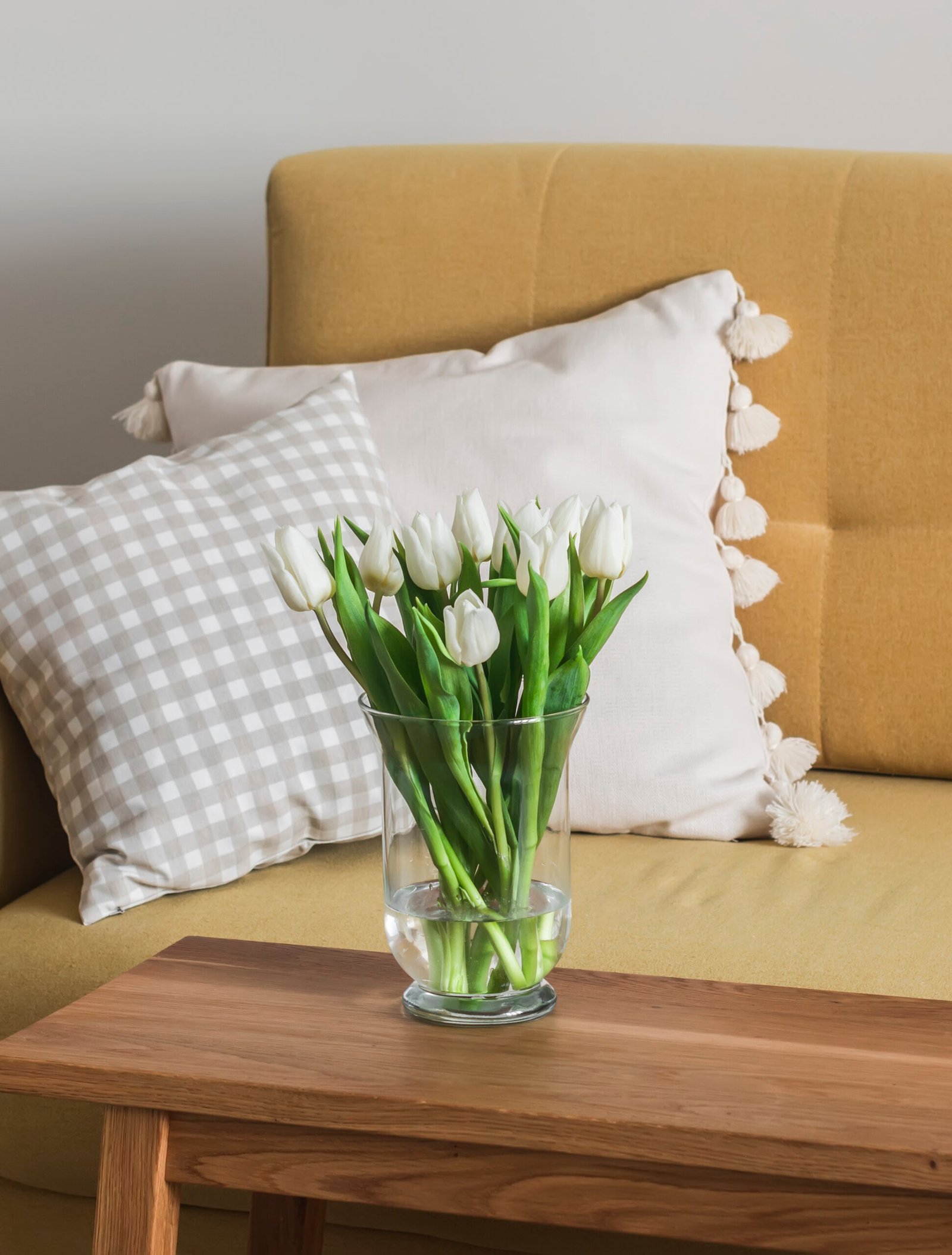 A bouquet of white tulips in a glass vase on a wooden table next to a yellow sofa in the living room