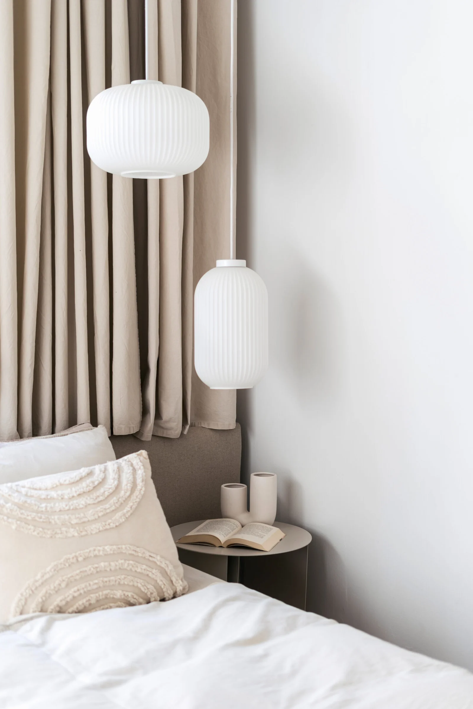 Detail of interior in cozy bedroom, open book and vase on night side table near comfort bed with bedding linen and cushions, modern pendant lamps and curtains in light tones. Home decor.