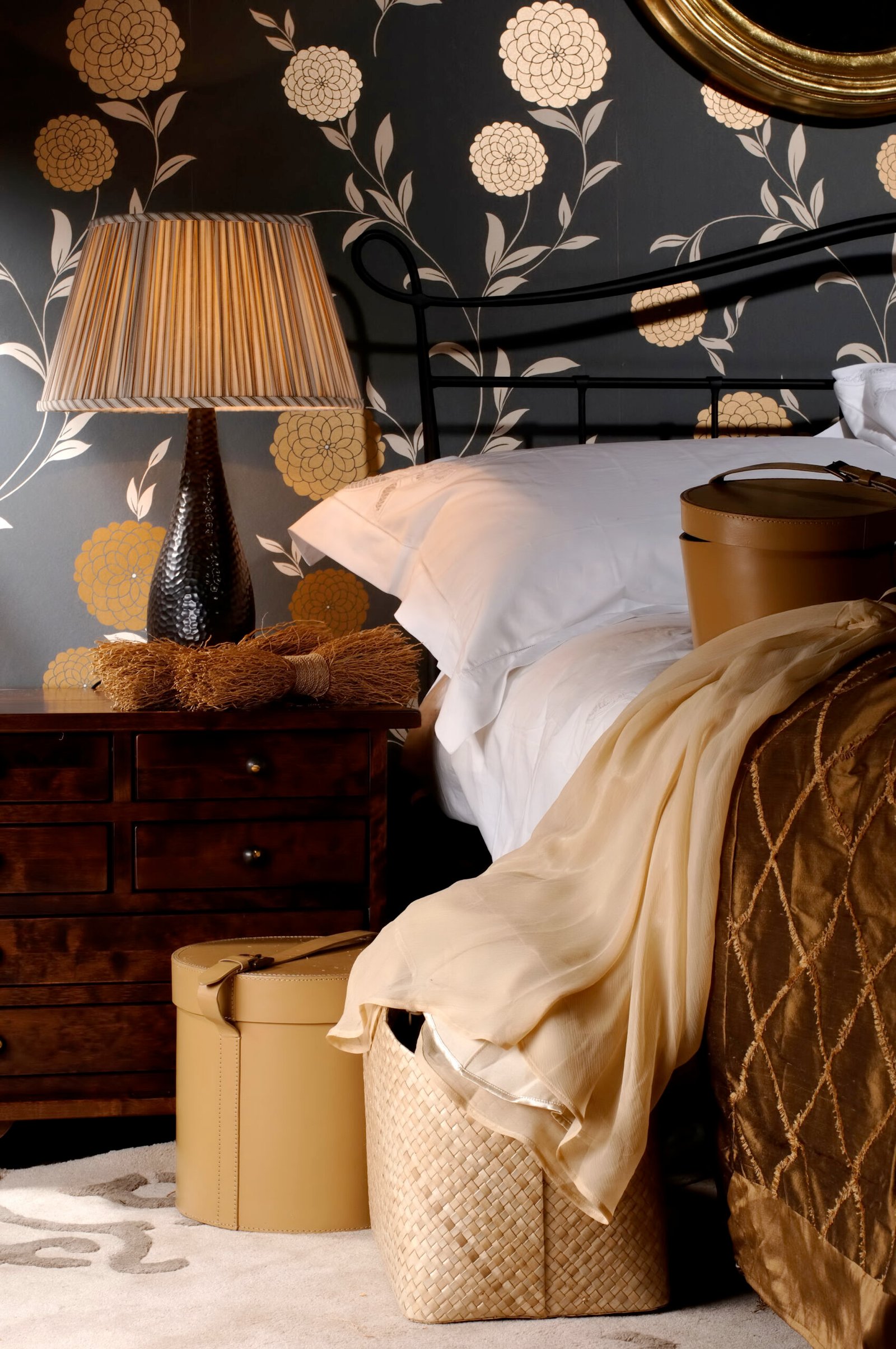 Fabric lampshade on the lamp at the big bed with pillows in the bedroom with floral print on the walls with suitcases