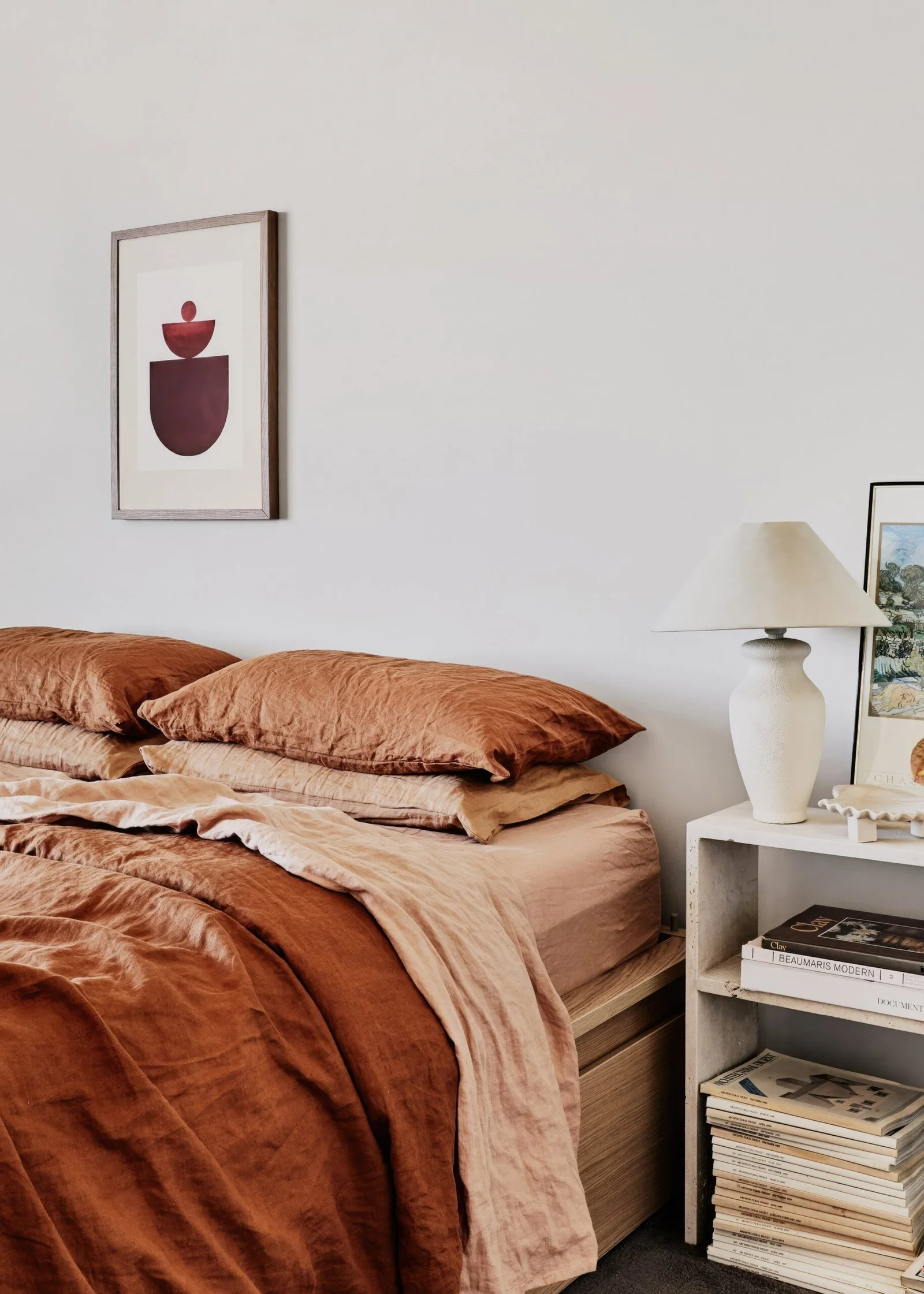 Rust bedding with a wall art above. White bedside table with a table lamp and decor pieces.