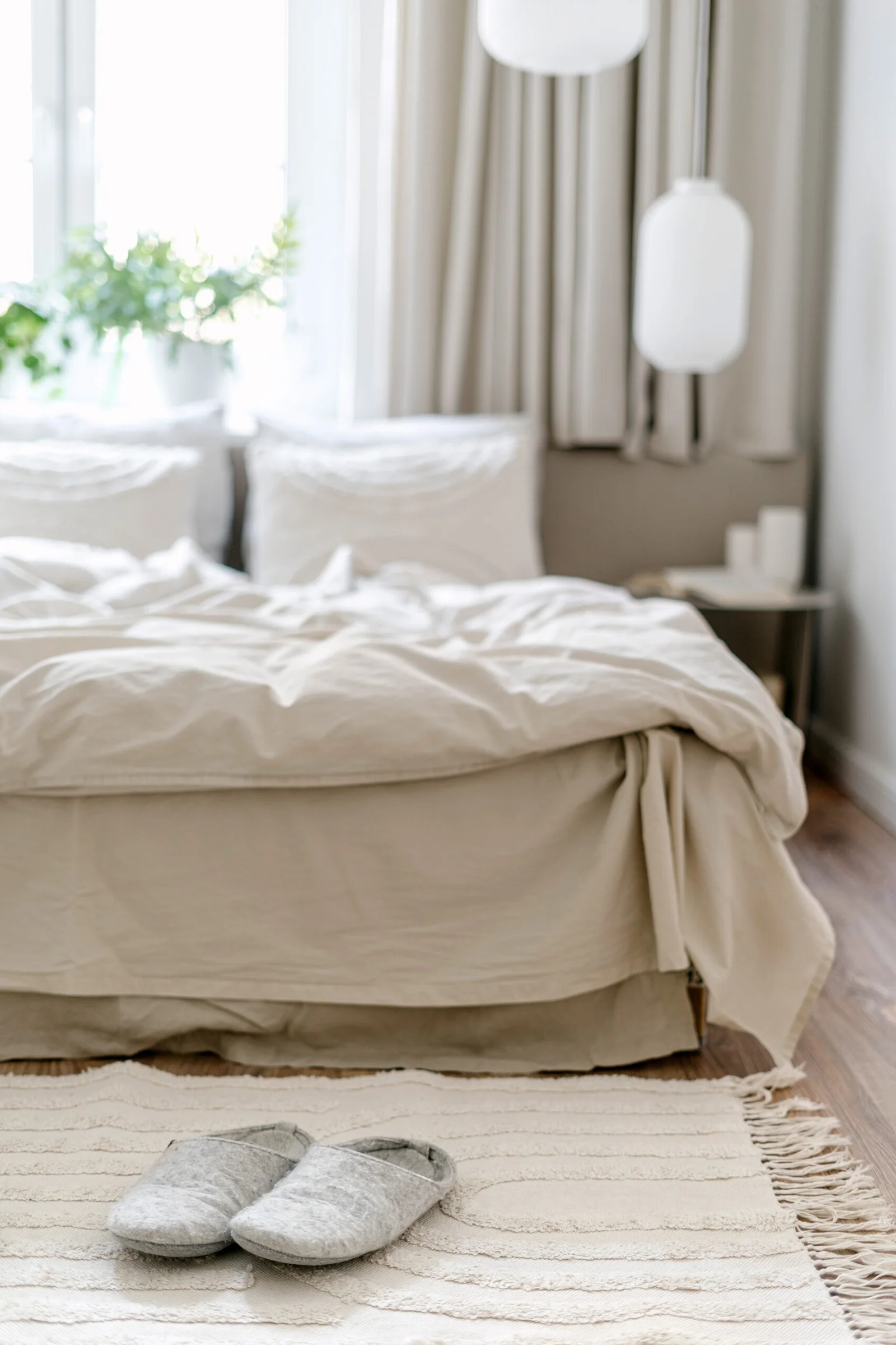 Selective focus on home slippers on floor carpet near comfort bed with pillows, blanket and bedding linen. Cozy bedroom with scandinavian interior in light tones. Hygge style in room