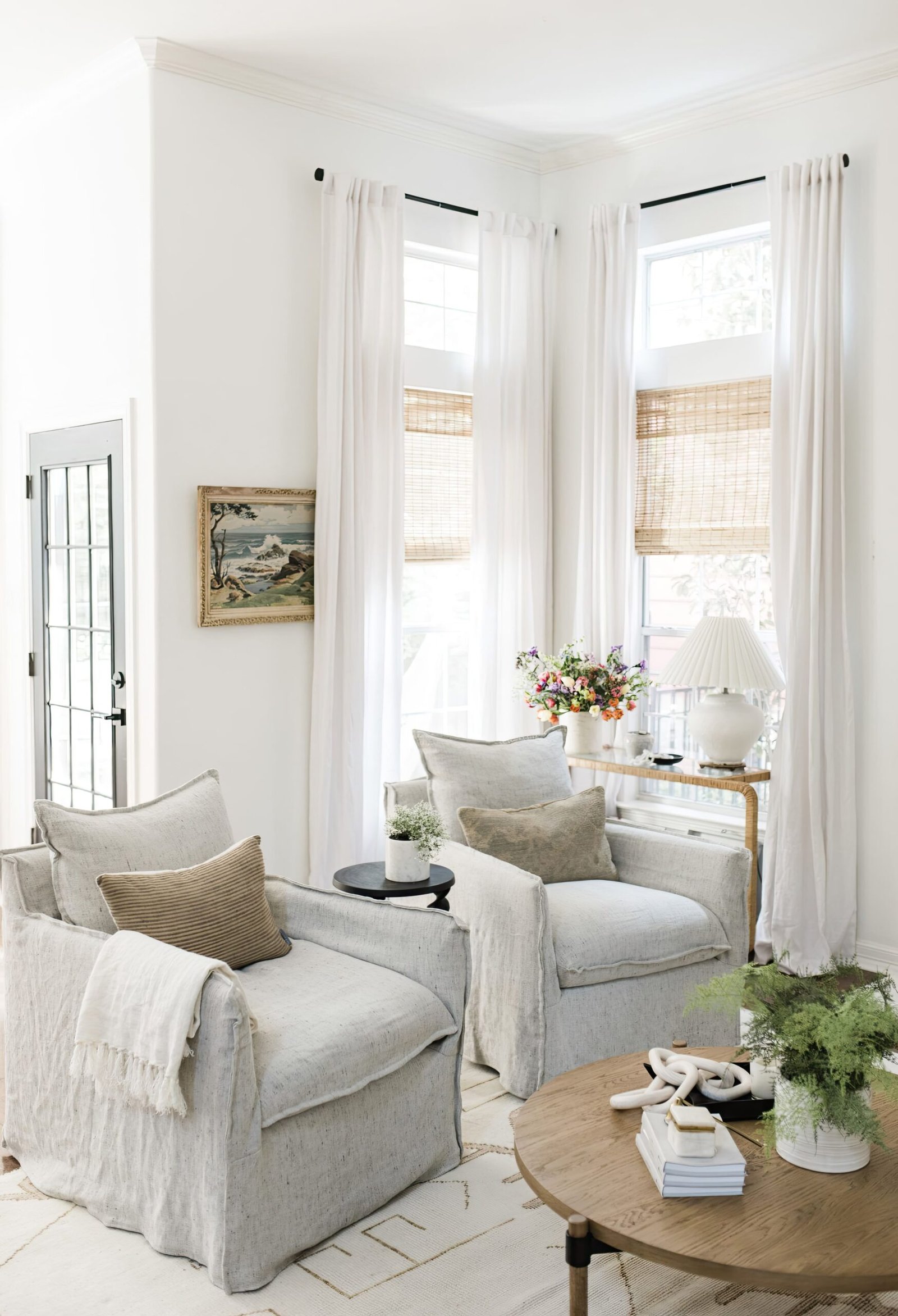 Living room with two light armchairs and a wooden coffee table. Behind is two large windows with tall curtains. 