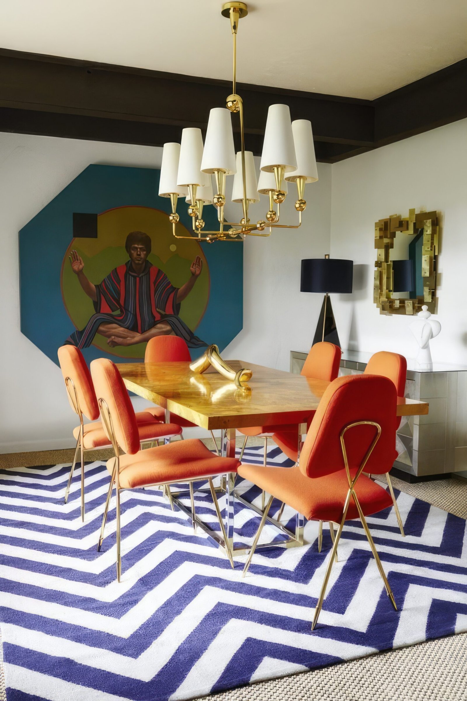 Eclectic dining room with geometric blue and white rug, a big wooden table and orange chairs. On the wall is a large wall art and above the table is a staple ceiling lamp. 