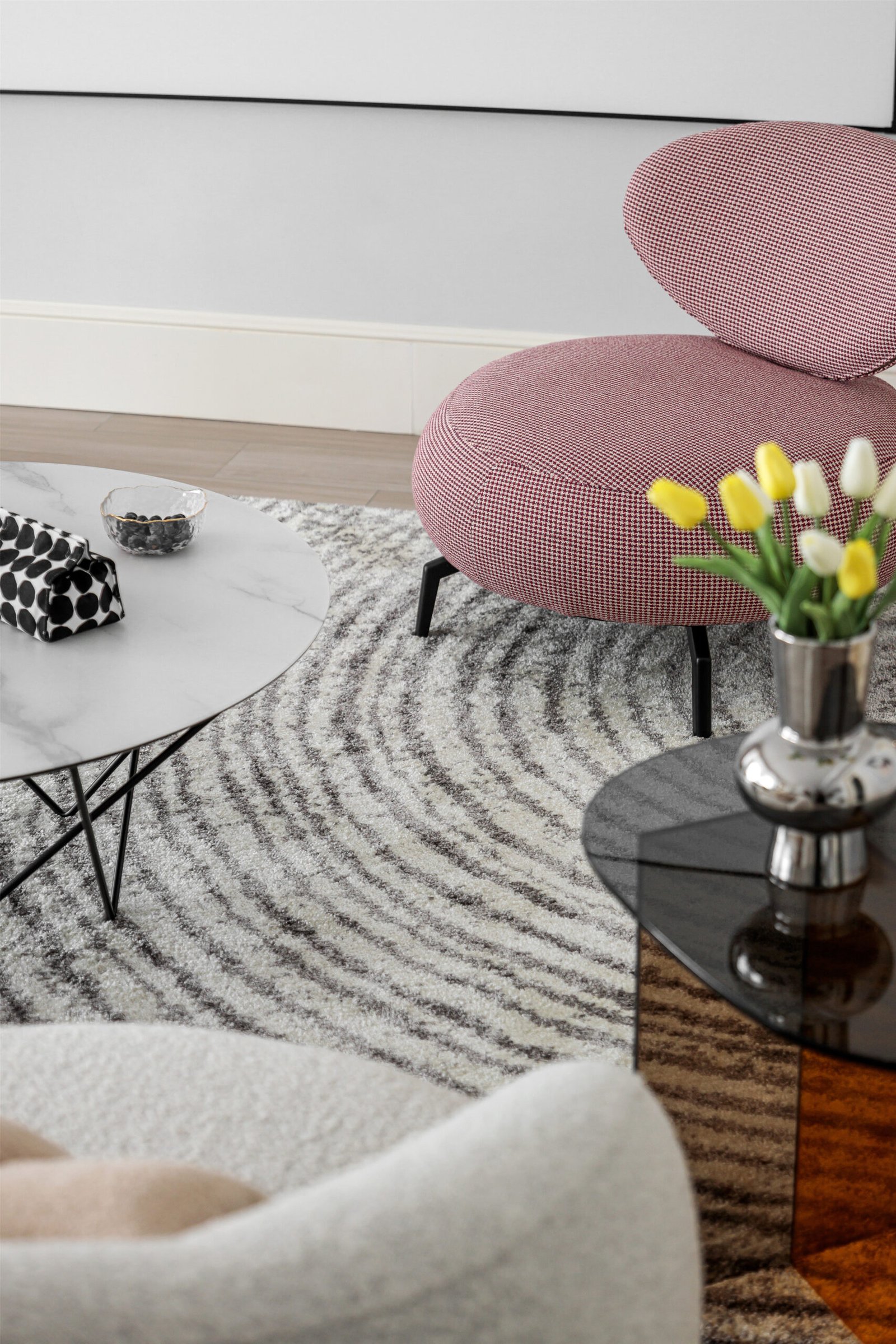 A closeup on a seating area with a pink armchair, a glass table, a vase with tulips and a marble table. On the floor is a large geometric rug.  