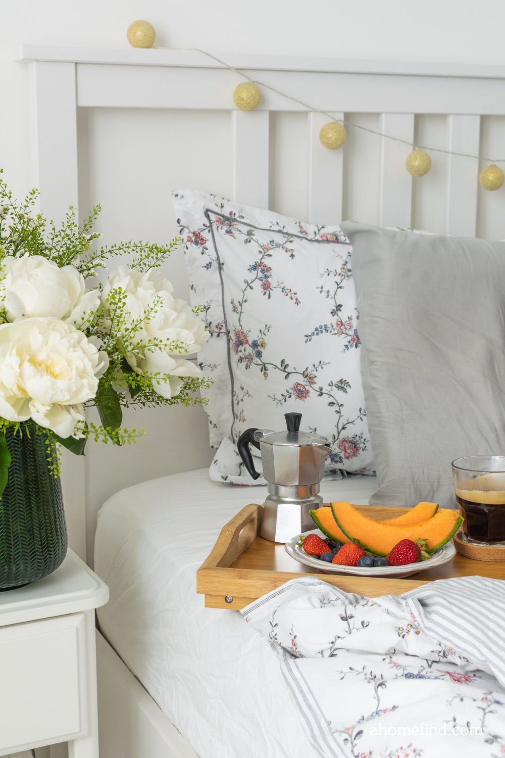 Floral print perfect bed sheets with breakfast on the bed. A white bedside table with a vase with flowers. 