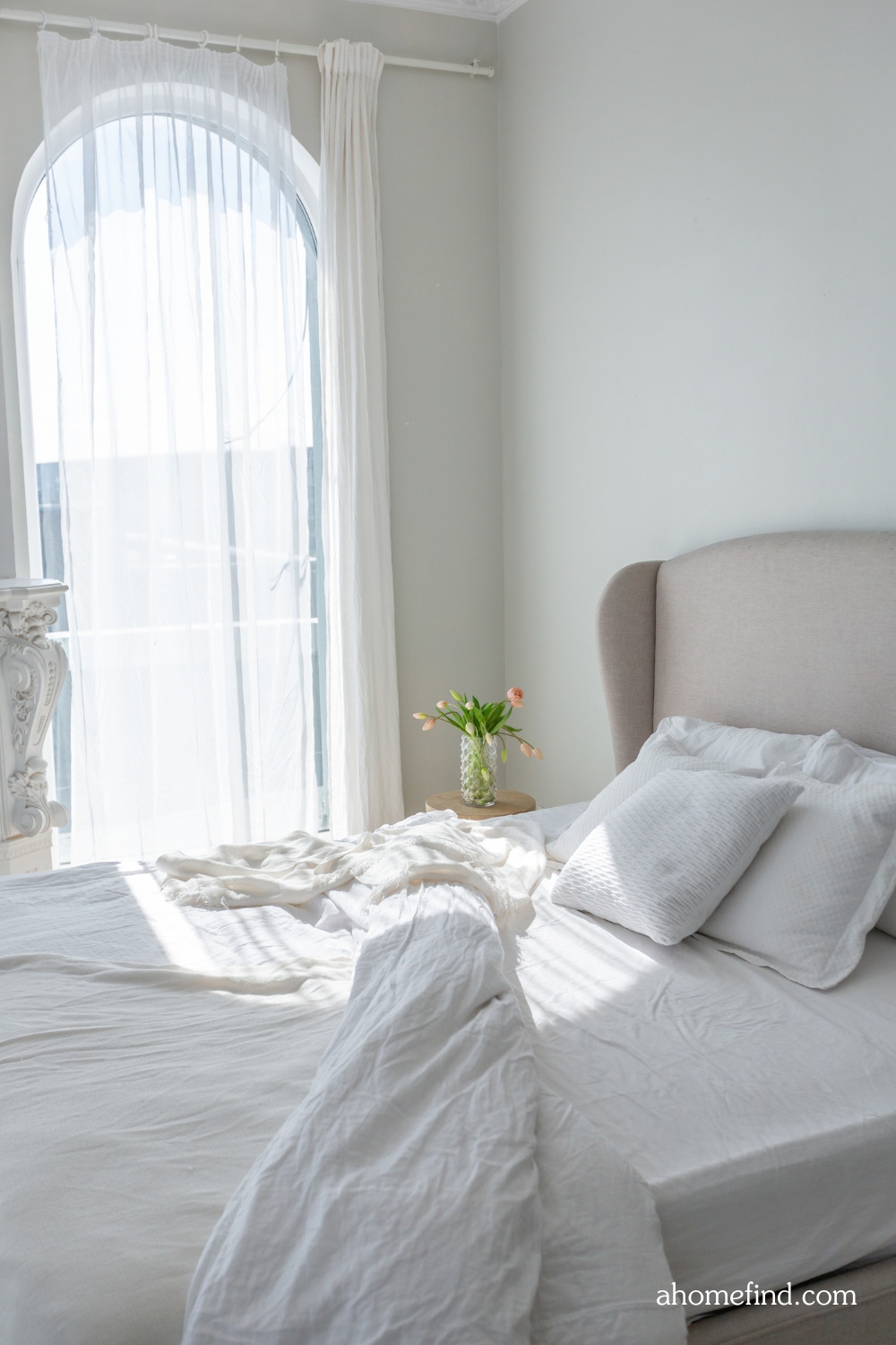 White bed sheets on a neutral bed in front of a window with white curtains. 