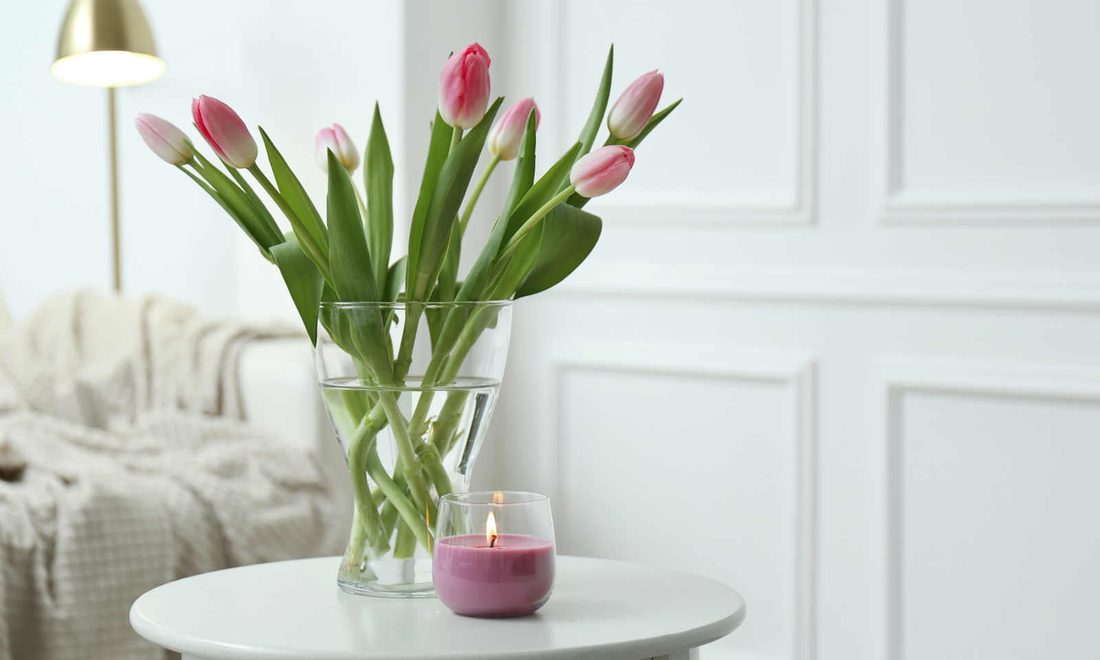 Spring tulips on a side table and a burning candle. In the background is a sofa. Scaled. Spring Home Decor.