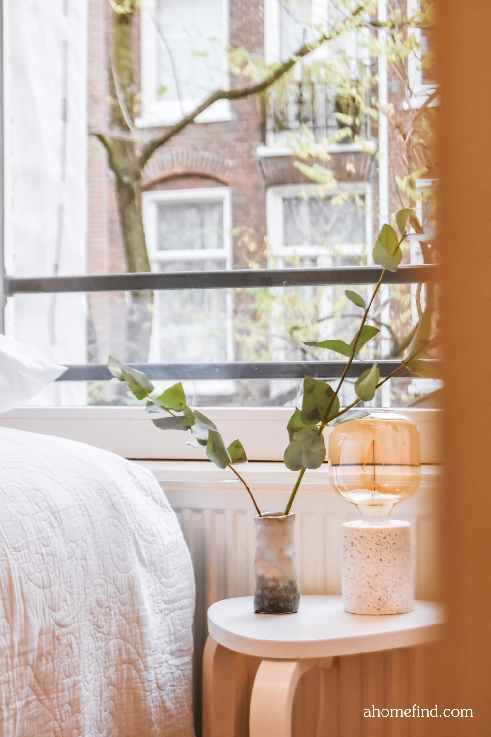 Small bedroom lighting tips with a table lamp and a vase with eukalyptus in front of window and bed. 