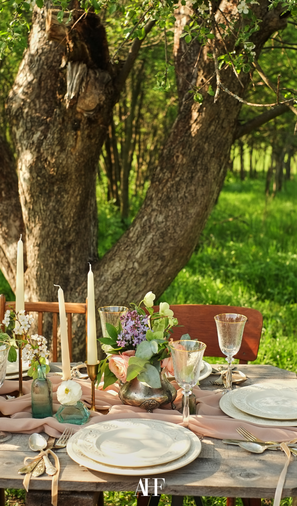Swedish midsummer table setting outside in the nature in front of a huge tree. Candlesticks and flowers on the table. Midsummer table decor.