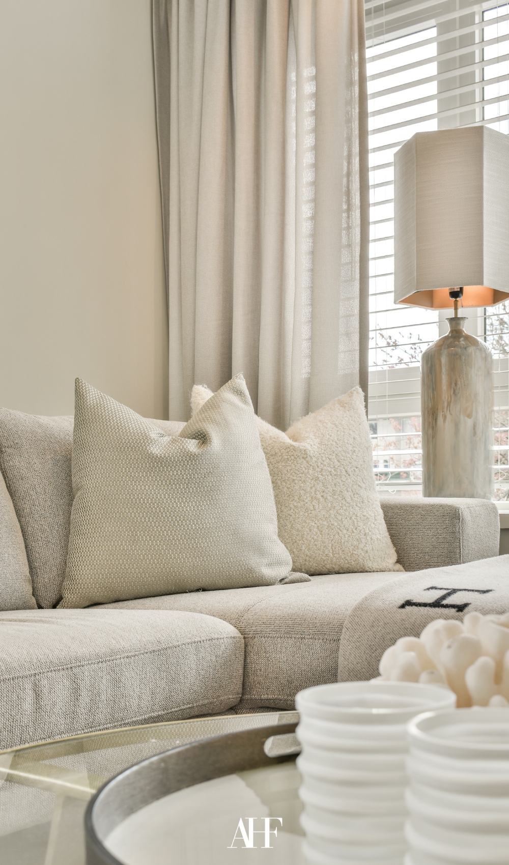 A cozy, light-filled living room featuring a white or ivory sofa, sandy beige throw pillows, sheer linen curtains. Summer Interior Design Trends.
