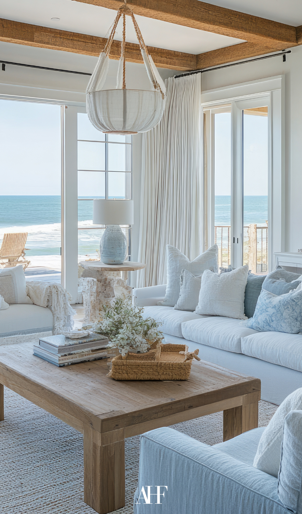A coastal living room with white sofas and a wooden coffee table, large windows towards the ocean.
