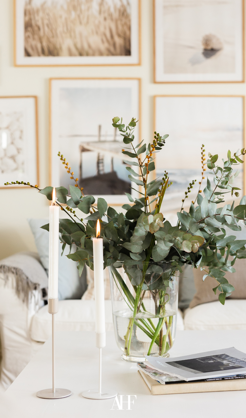 A cozy living room with a closeup on a coffee table with greenery in a glass vase, plus two candle holders and wall art on the wall.