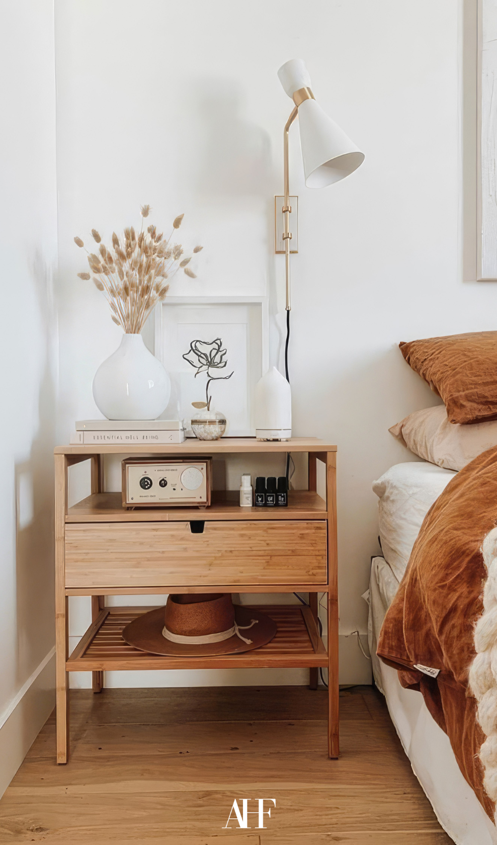 A bedside table in wood with lots of decorations on it. To the right is a bed with rust bedding.