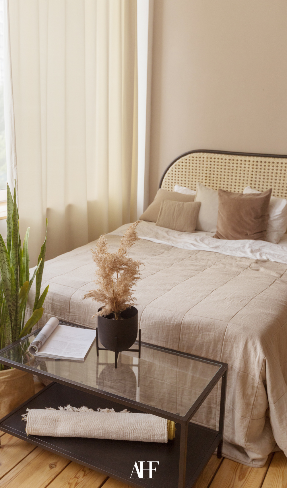 A cozy bedroom with earthy tones and a rattan bedframe, beige bedidng and a large window.