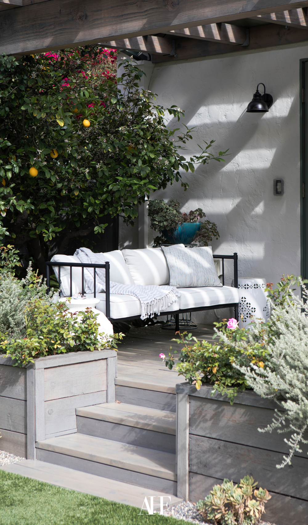 A black couch with bright white pillows, lots of plants and a wooden deck.