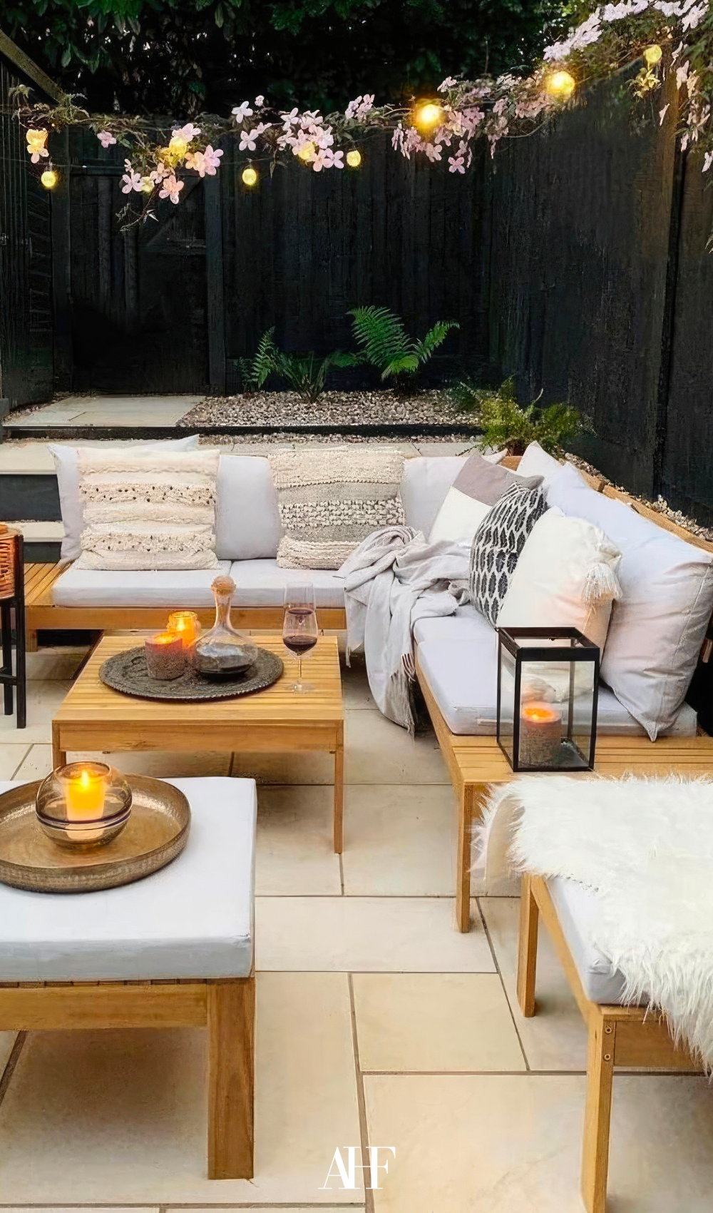 A large lounge set in wood with light cushions and pillows. Cozy lighting and plants in the background.