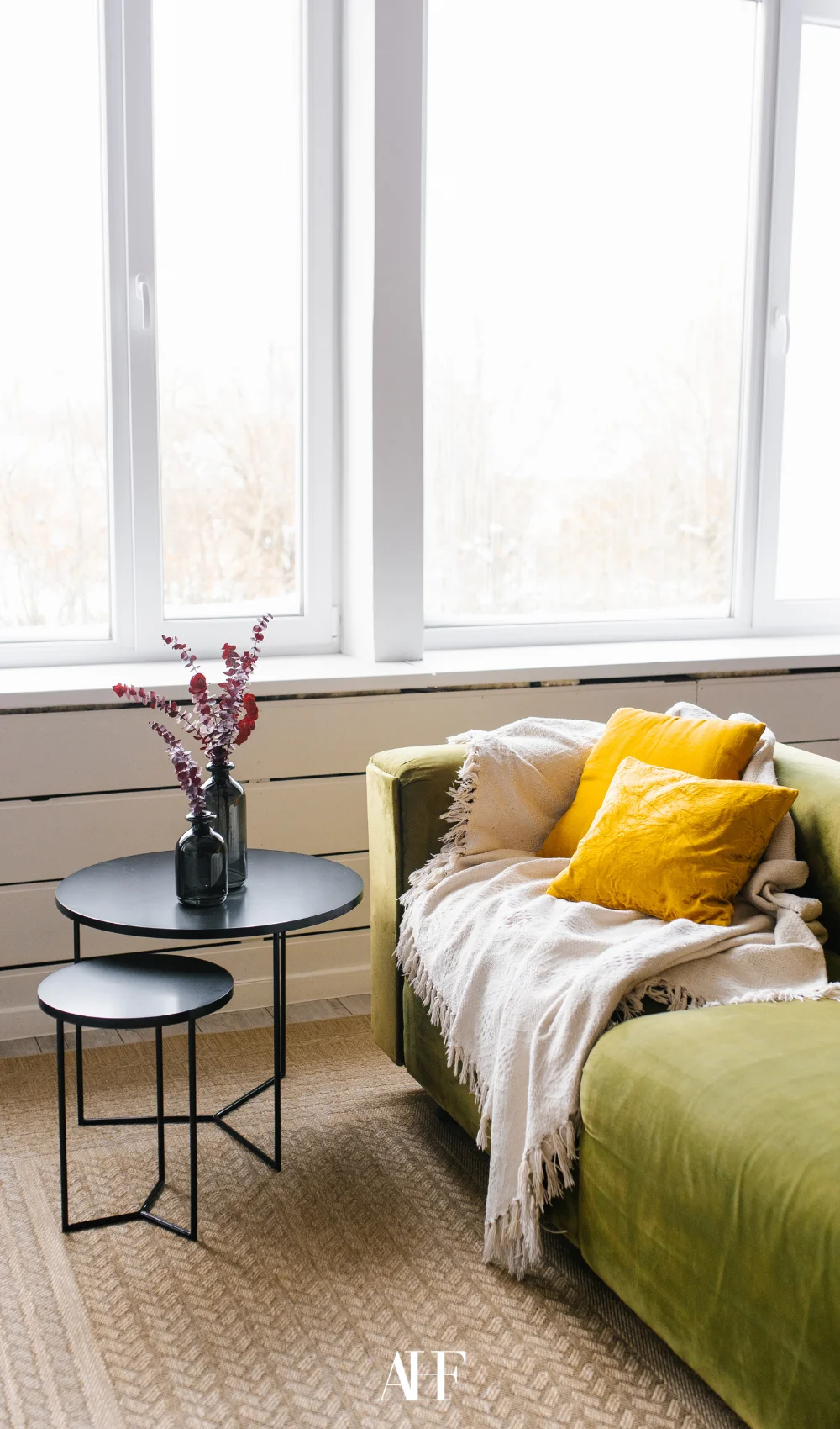 A green sofa with strong yellow pillows, small black coffee tables and a large window behind.