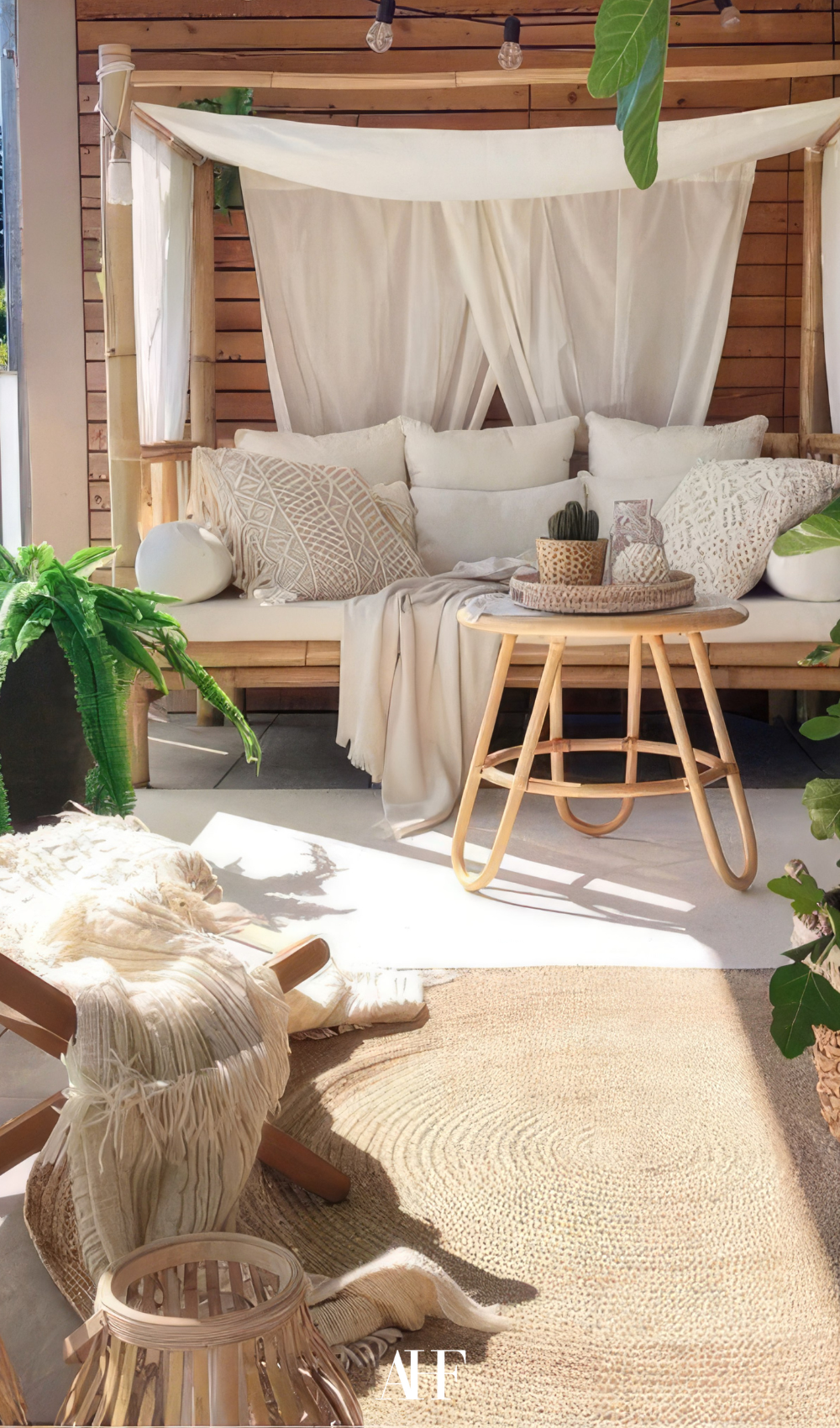 A cozy boho small summer patio with a wooden couch with white pillows and blankets. A rattan armchair and a bamboo coffee table. A white rug and some plants.