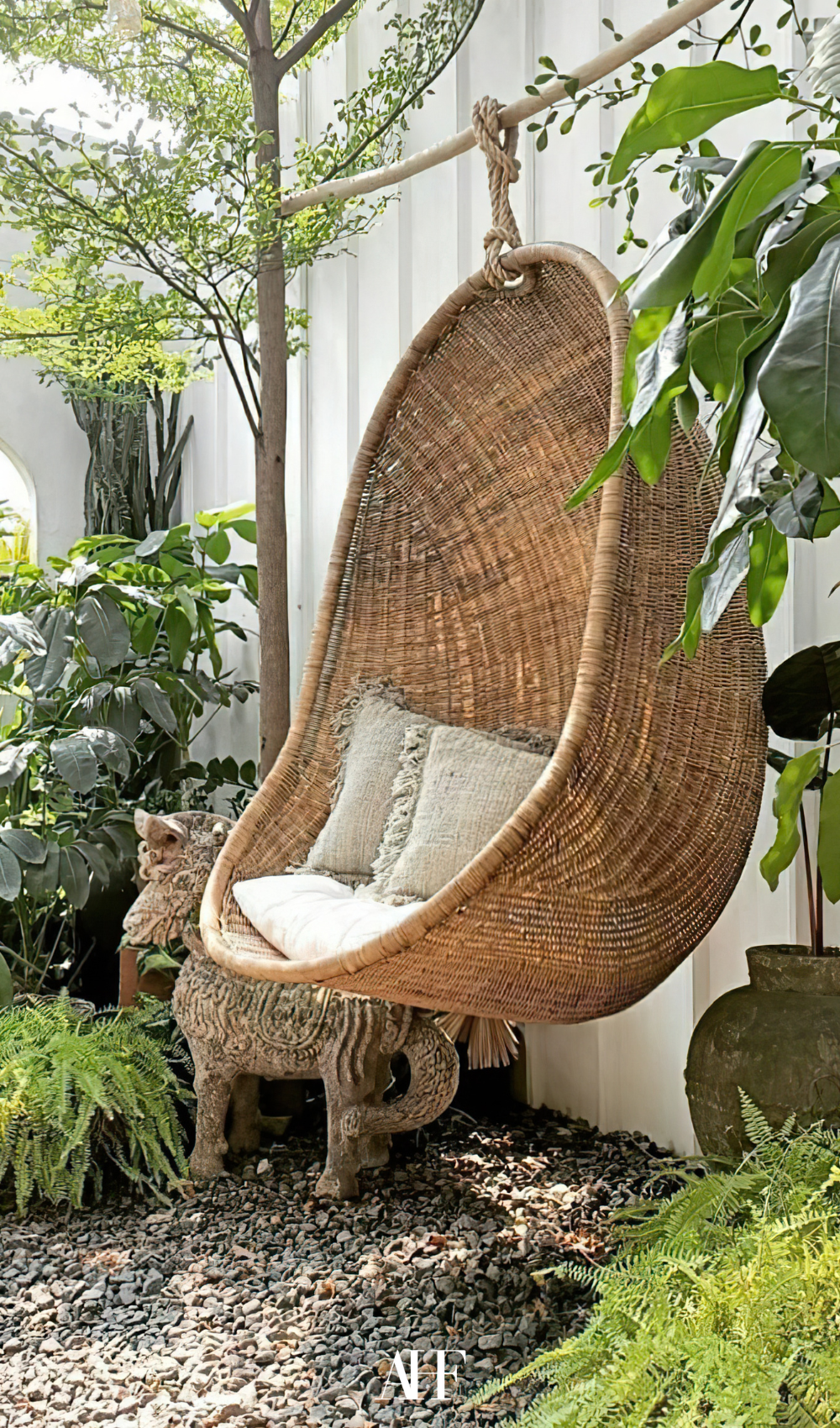 A large egg hammock in rattan material, some white pillows inside and lots of plants.