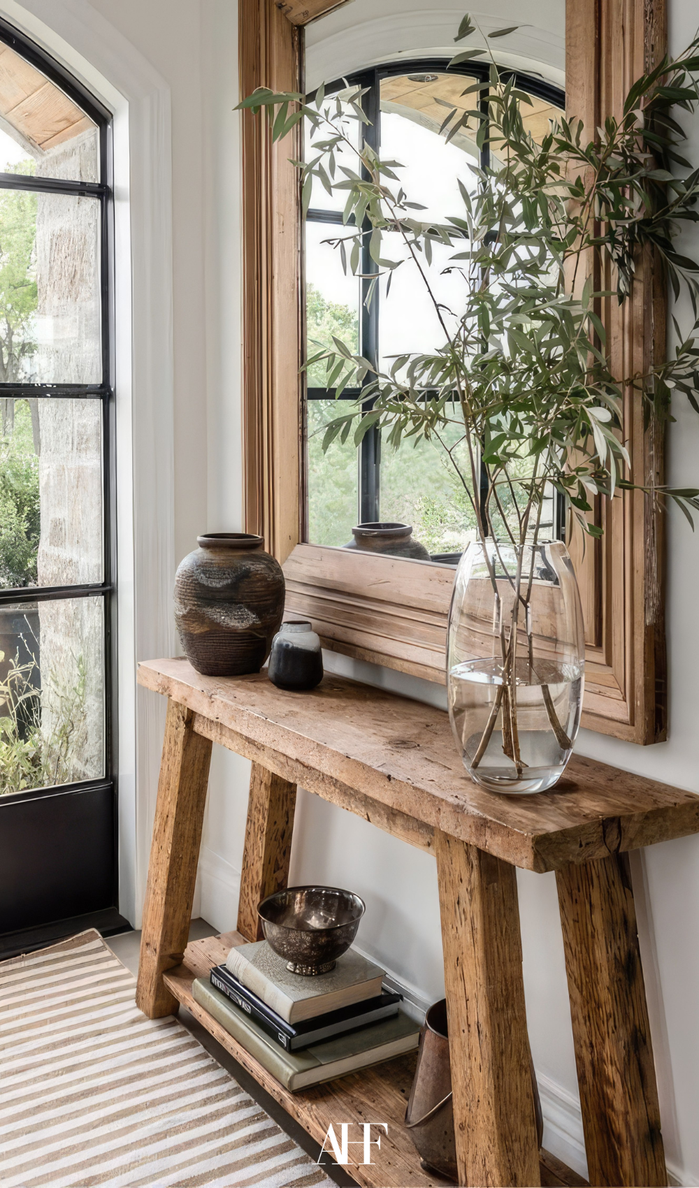 A rustic wooden side board bench with a large mirror on the wall, plus a clear glass vase with plants in it.