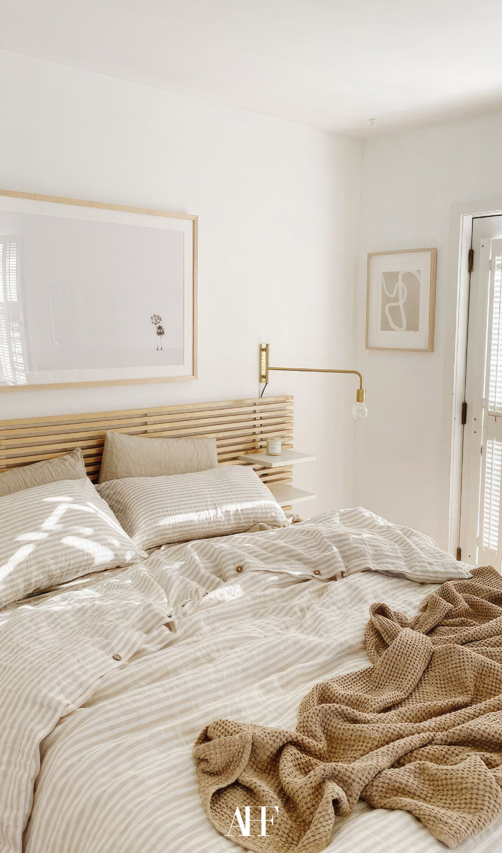 A wooden bed frame and beige bedding, white walls and a wall art.