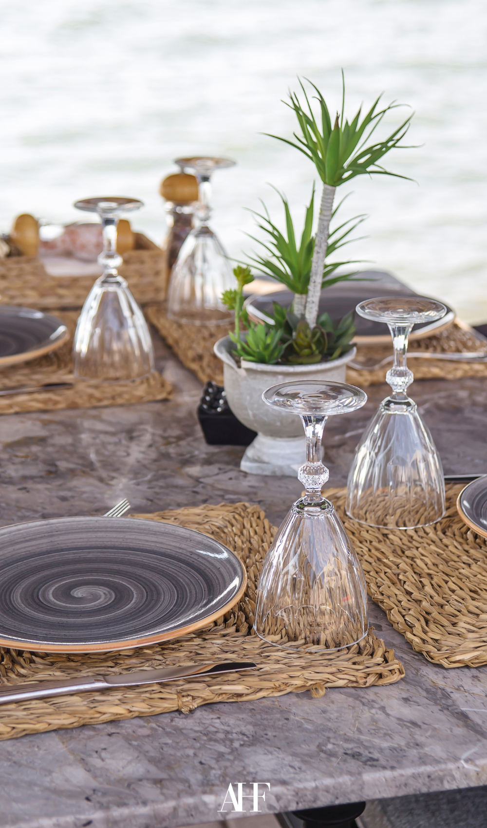 Summer table settings with a wooden table, rattan coasters and grey plates. Glasses and a green centerpiece. Table by the ocean.