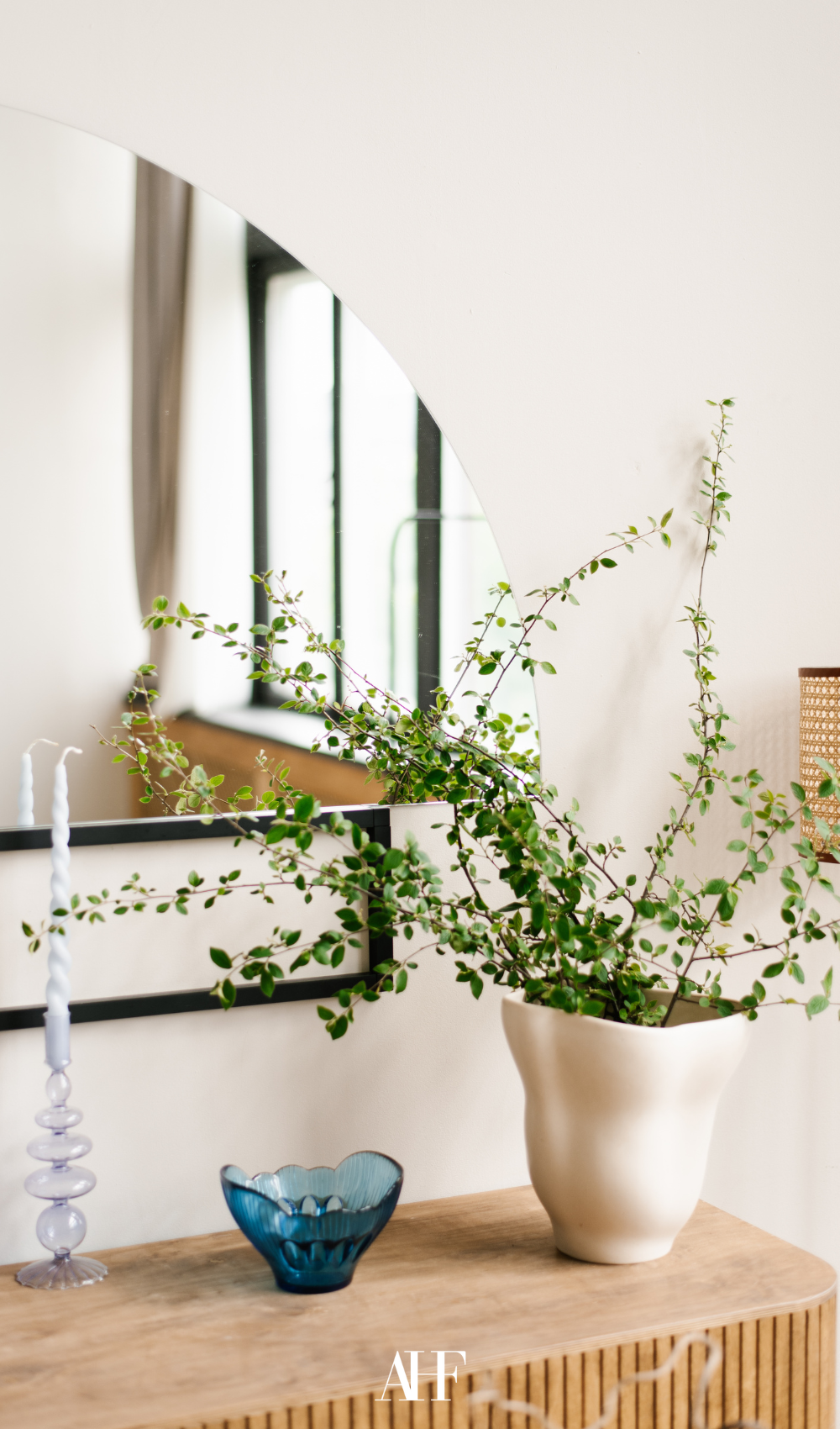 A wooden sideboard with a large mirror on the wall, a white planter with a green plant in it. Two candle holders.