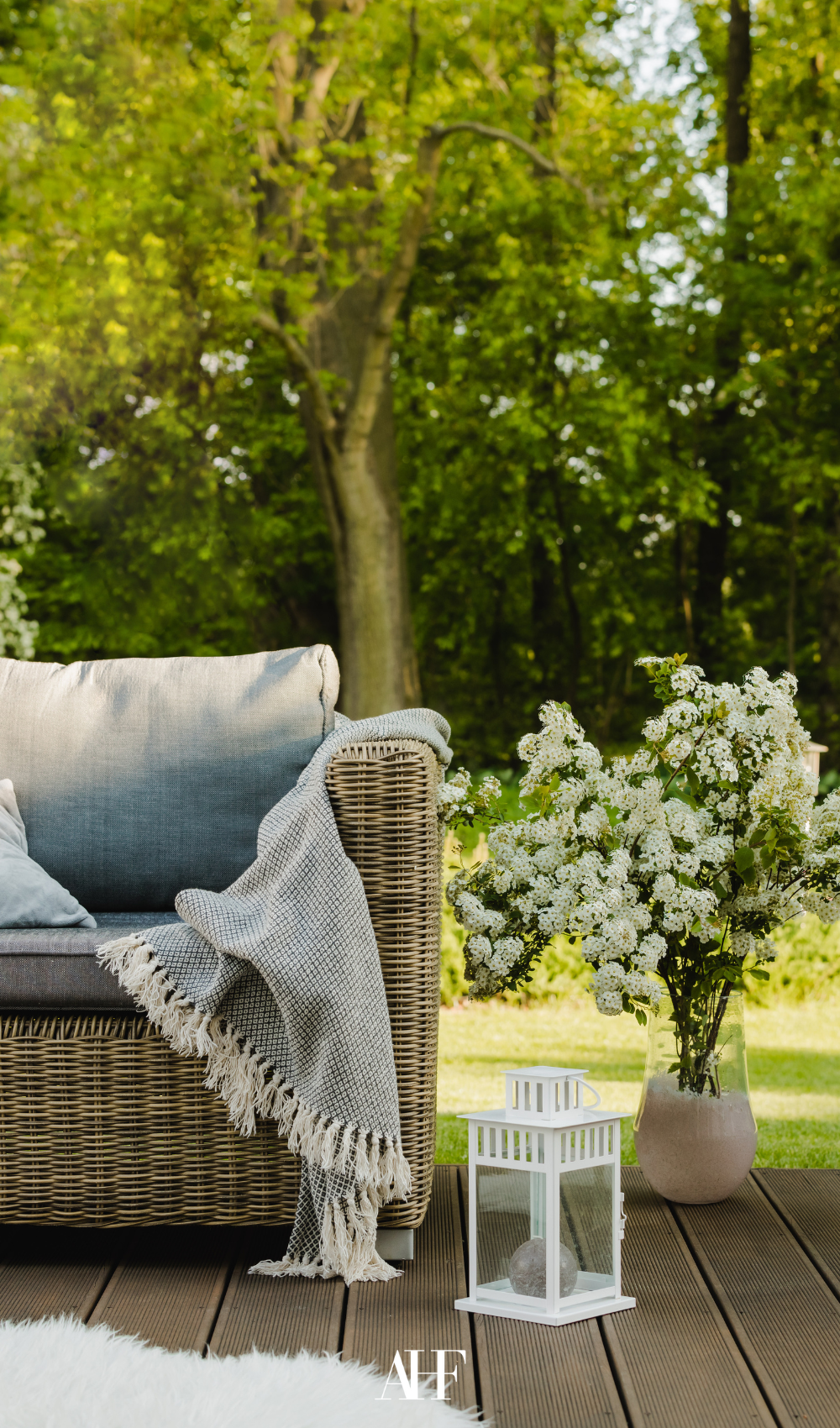 Rattan couch with a vase of flowers next to it, as well as a lantern. In the background is the woods.