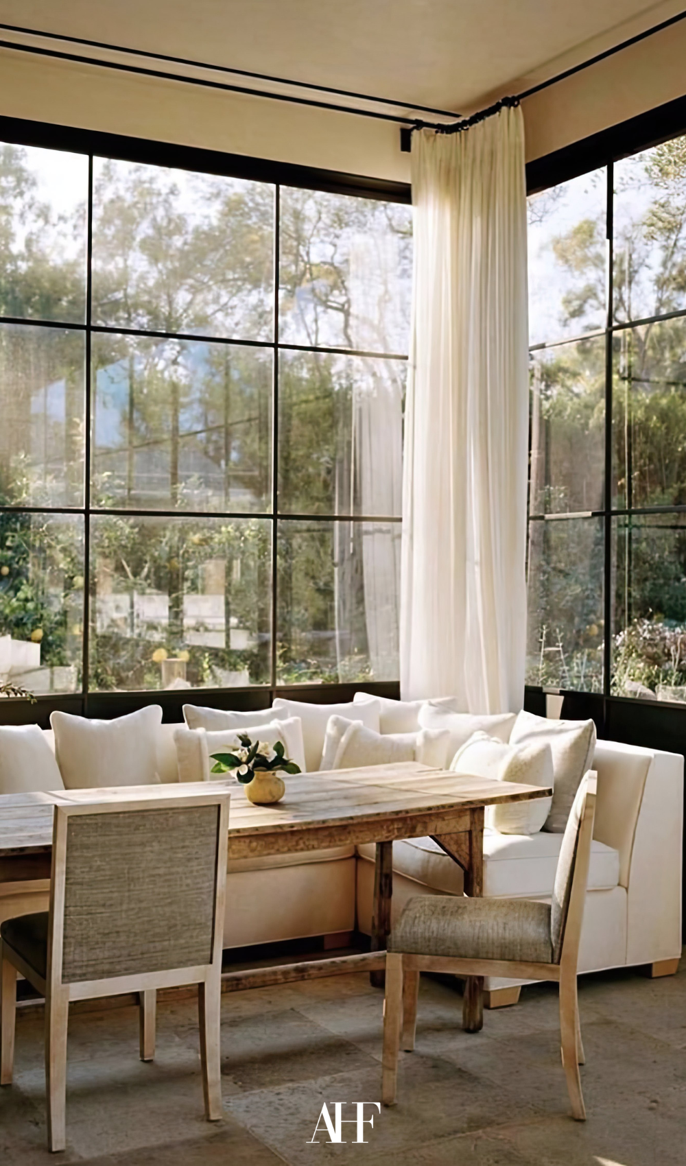 Wooden dining table with a white couch, two dining chairs and large windows. Sheer white curtains.