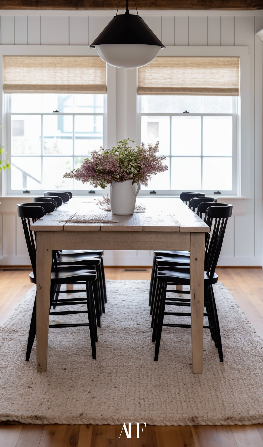 A wooden dining table with black chairs, a flower on the table. Two large windows, a perfect rug underneith the table. 