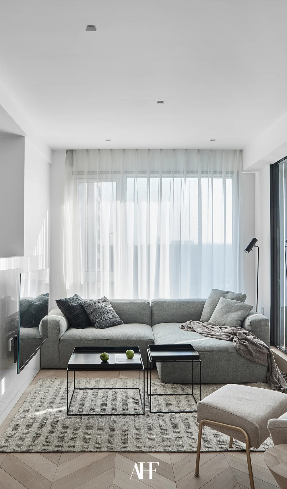 A grey sofa in front of large windows with see-through curtains. A TV on the wall and a black coffee table. Small apartment living room.