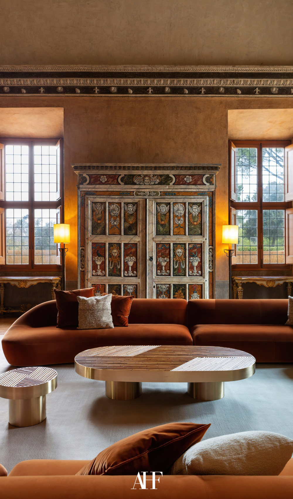 A rust colored beautiful sofa with a coffee table and traditional modern decor. Large windows.
