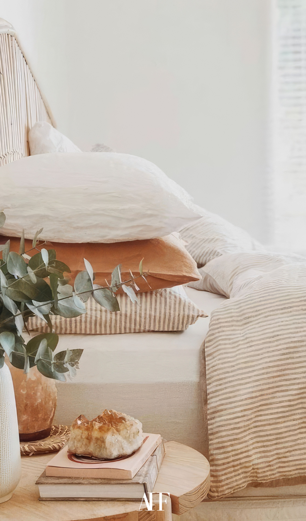 a cozy bed from the side wiht three pillows on top of each other and a wooden bedside table with a plant.