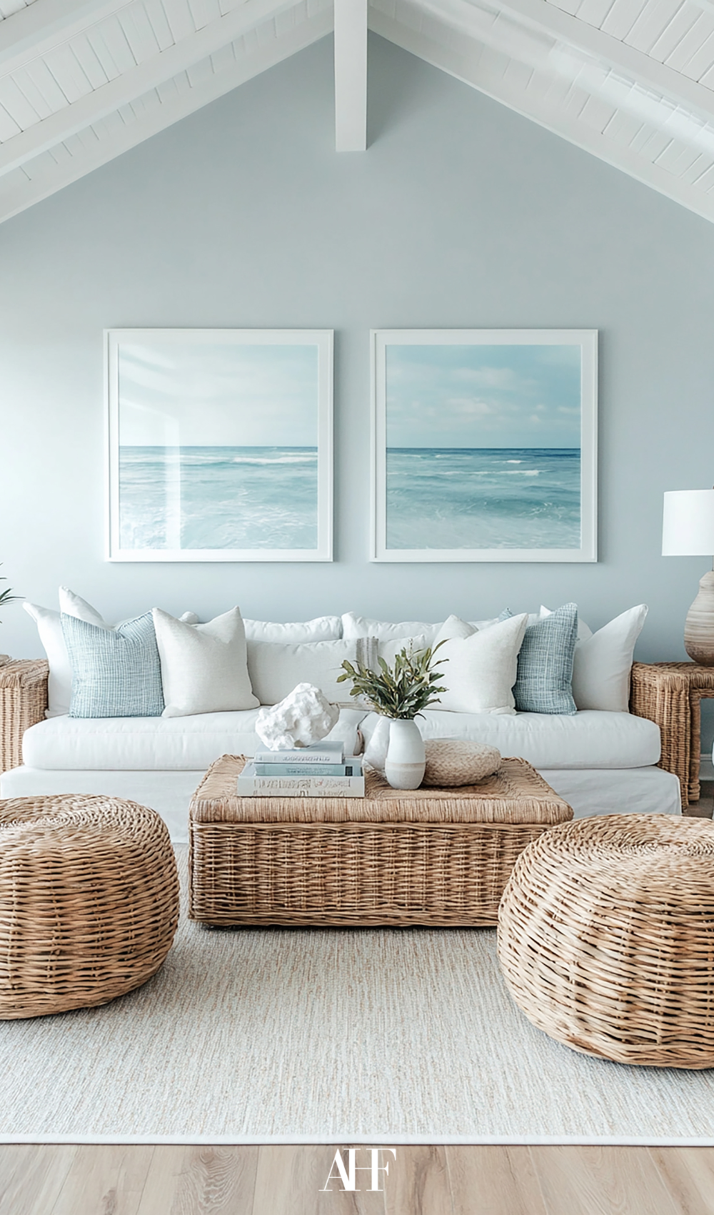A coastal blue living room, with the coast on wall art and a white sofa with blue pillows. Rattan coffee table and footstools.